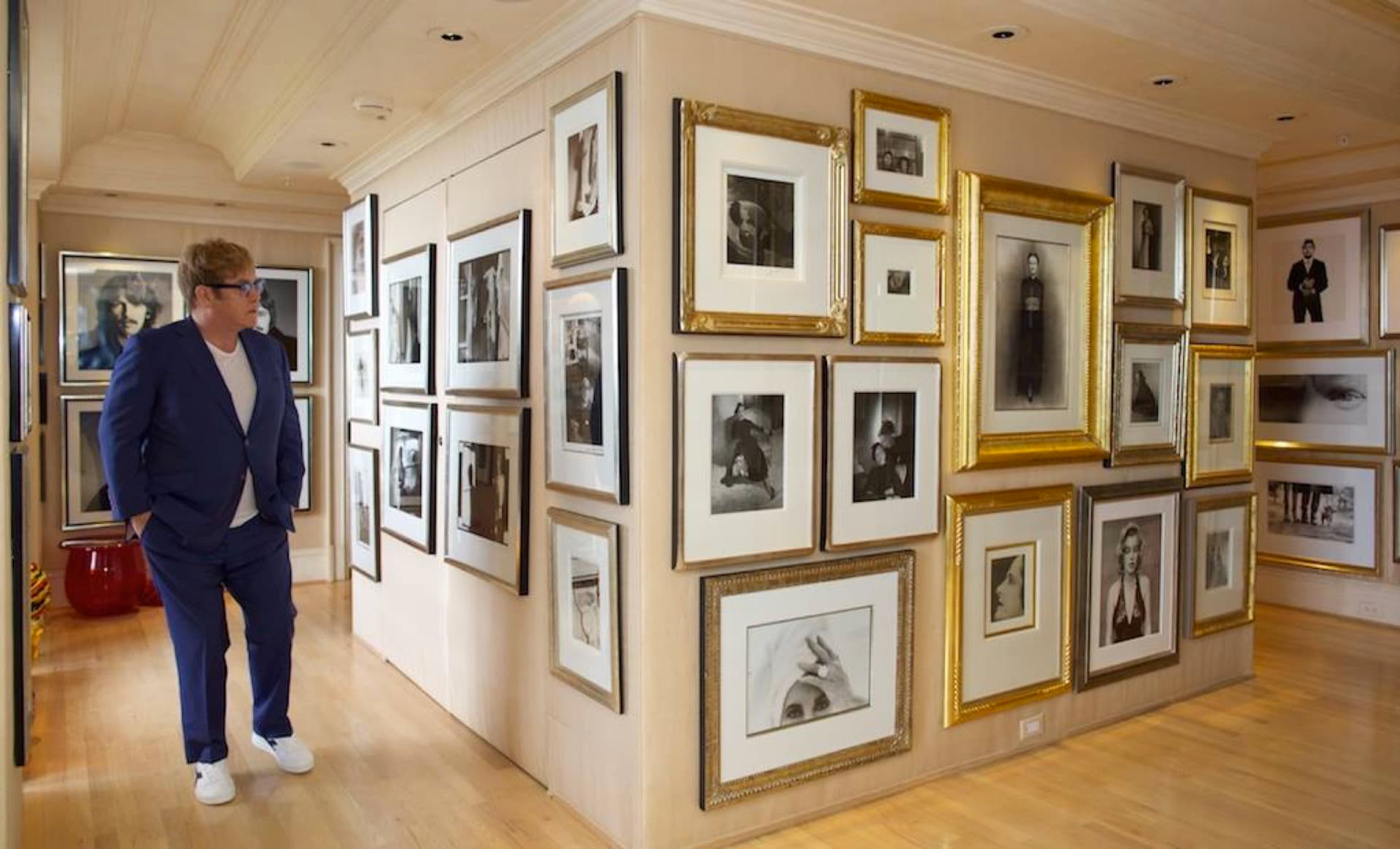 An image of Elton John in his house, surrounded by some of his photographs hanging on the walls.