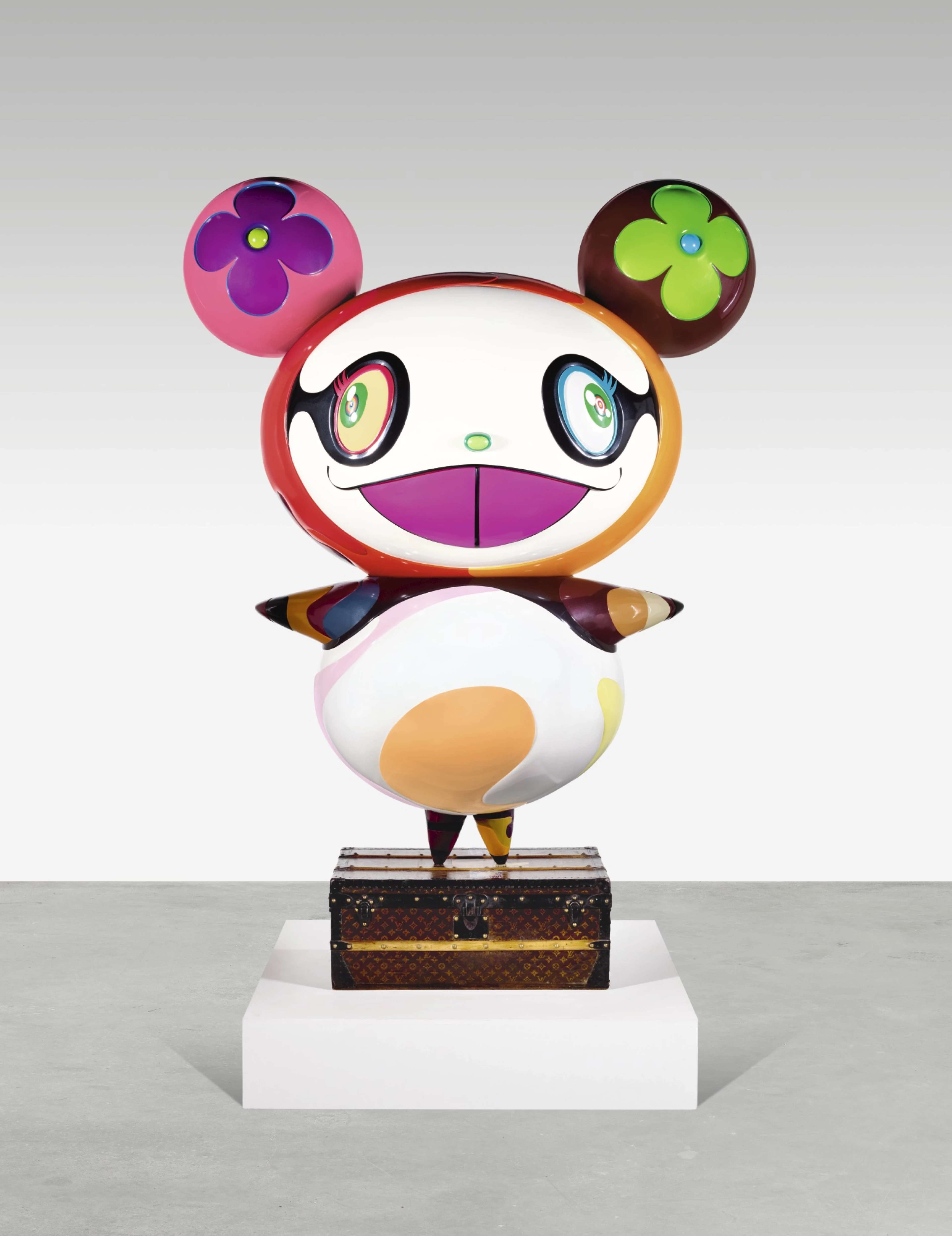 A colourful sculpture of a cartoon-like panda figure by Takashi Murakami, featuring a round body with multicoloured patches and large, expressive eyes. The character has oversized ears adorned with Murakami’s signature flower motifs, one pink with a purple flower and the other brown with a green flower. The figure stands on a Louis Vuitton trunk, placed on a white pedestal in a gallery setting.