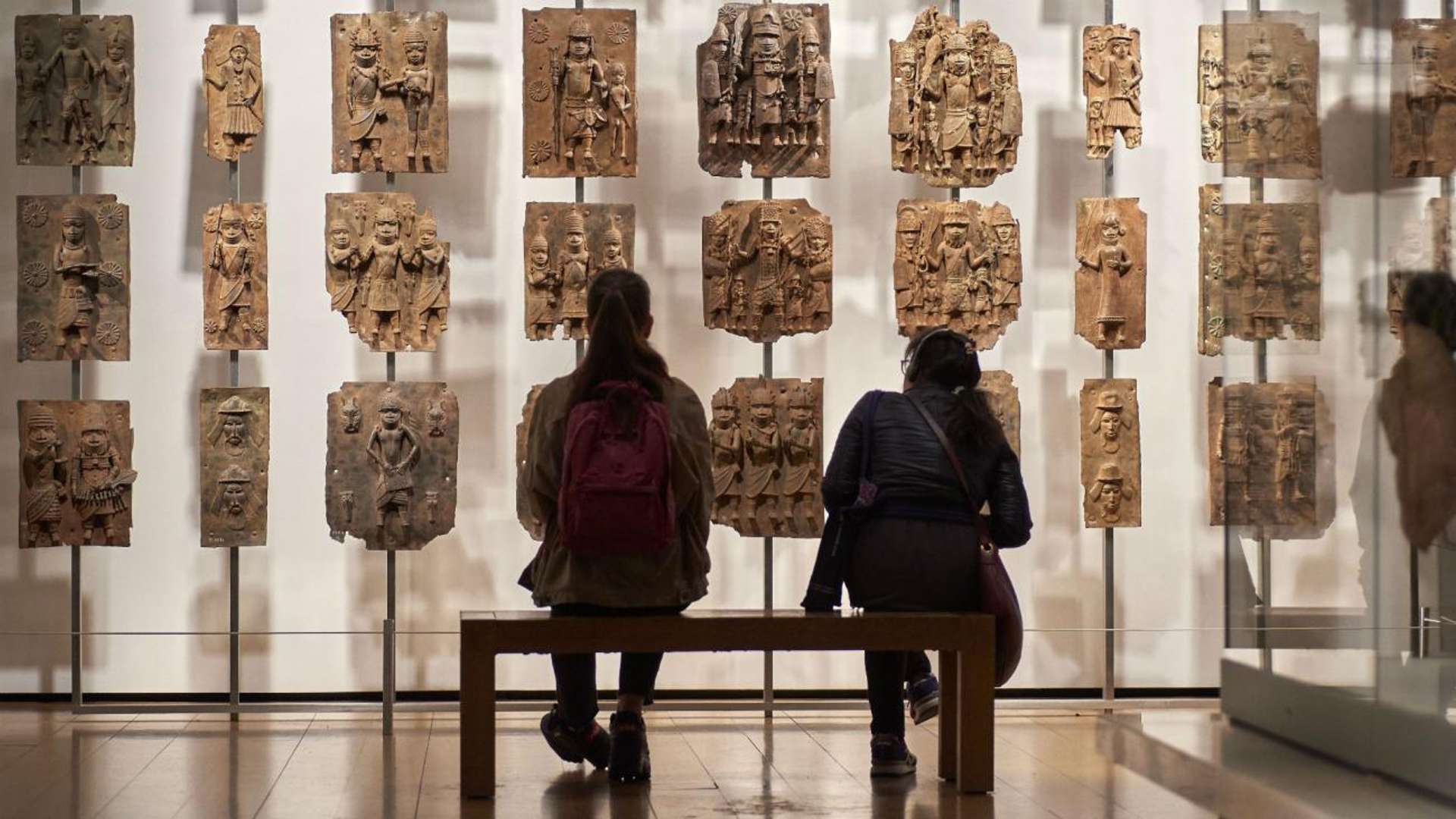 An image of two visitors sitting in front of the Benin Bronzes on display at the British Museum.
