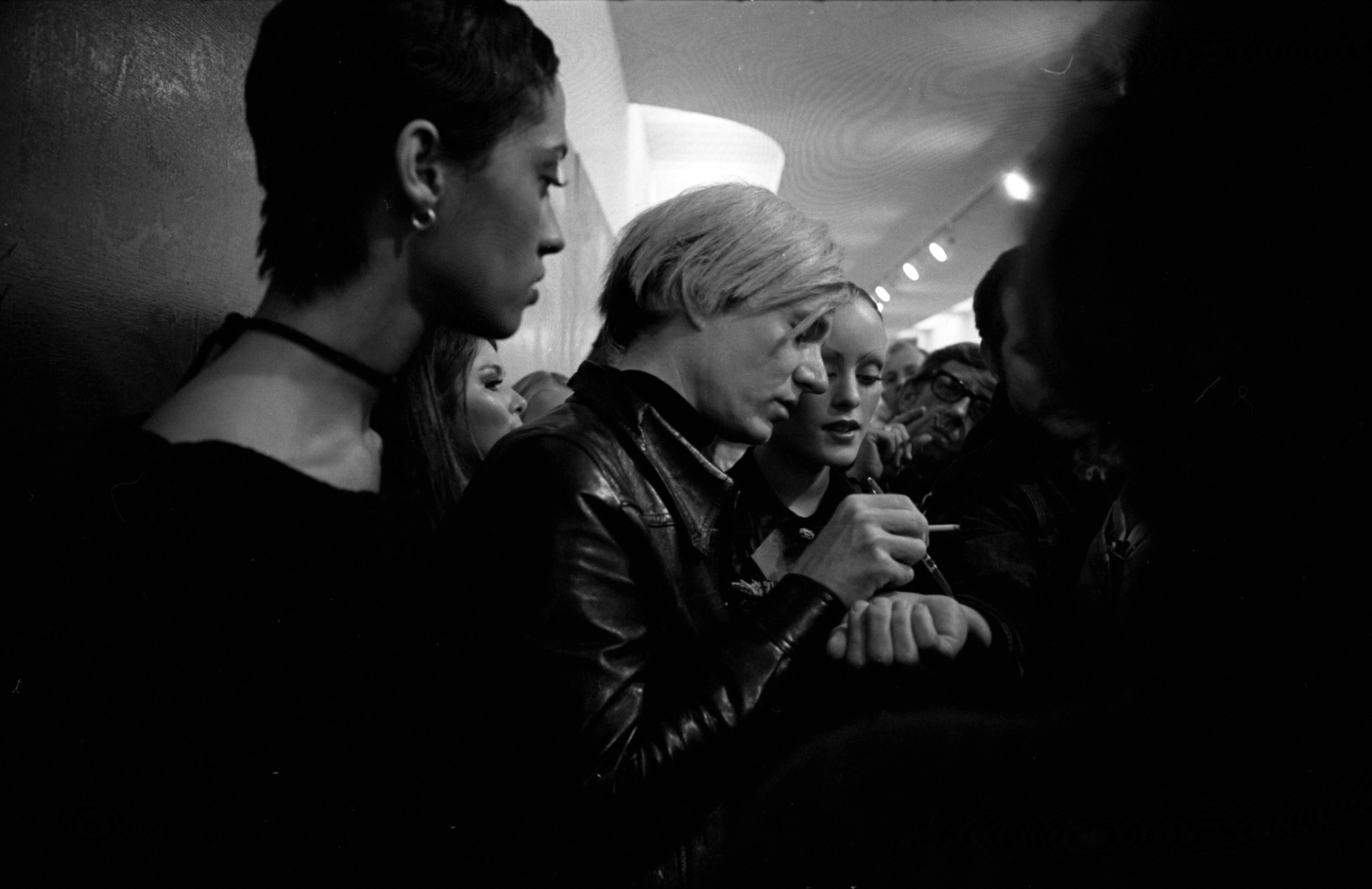 A monochrome image of Andy Warhol, surrounded by people, signing autographs and smoking a cigarette. He is wearing a black leather jacket.
