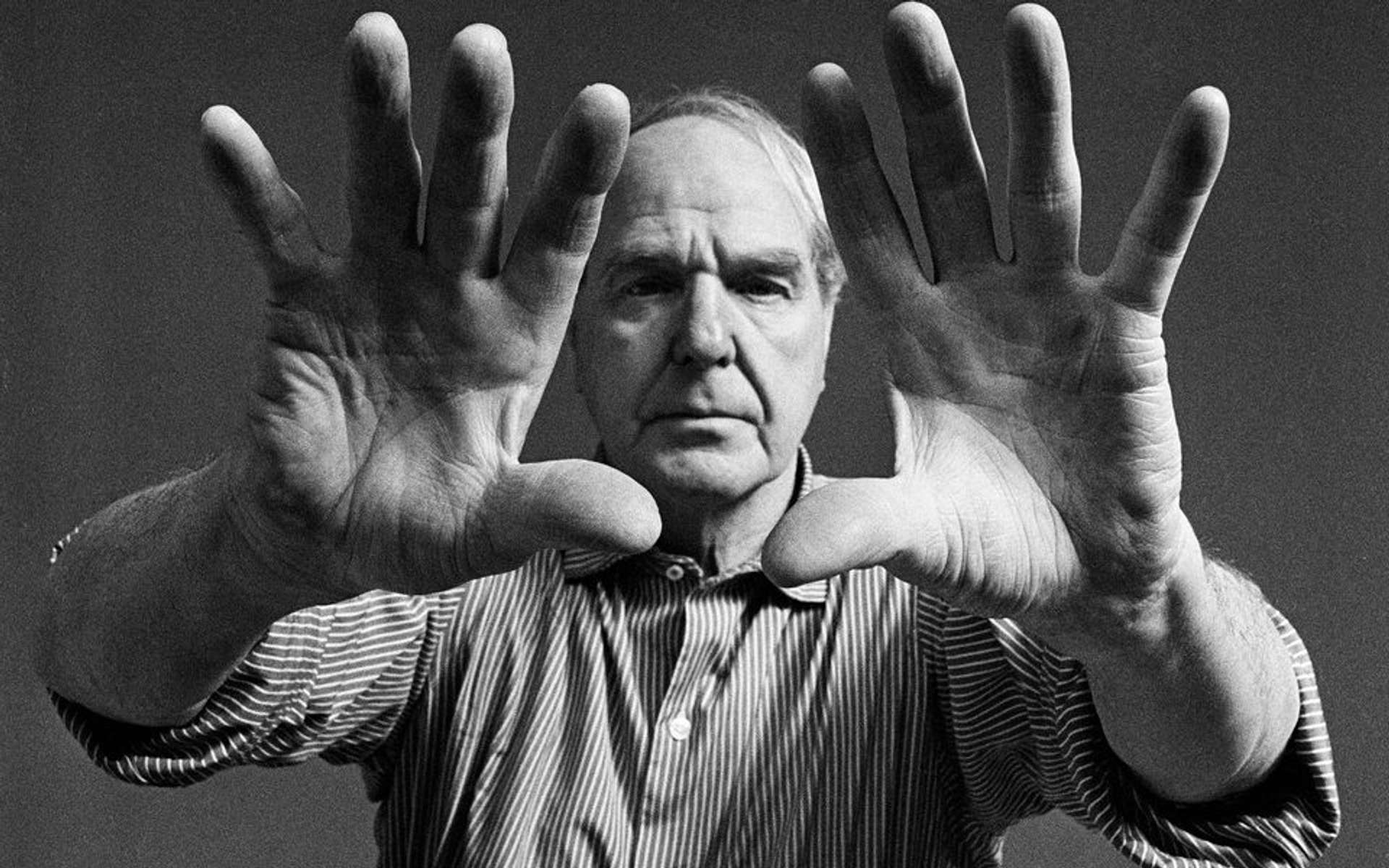 Black-and-white photograph of Henry Moore wearing a striped button-up shirt, with rolled-up sleeves. His hands, with fingers spread open, are positioned close to the camera lens, revealing the full frontal view of his palms.