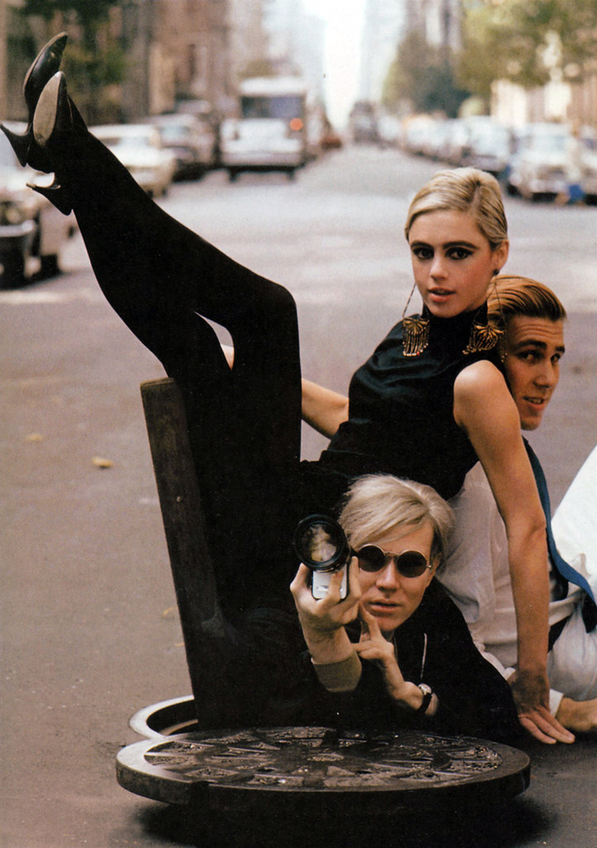 In this photograph, Andy Warhol is seen emerging from a sewer grating while holding a camera. Muse Edie Sedgwick is sitting over him, extending her long legs by her side and leaning against Chuck Wein.