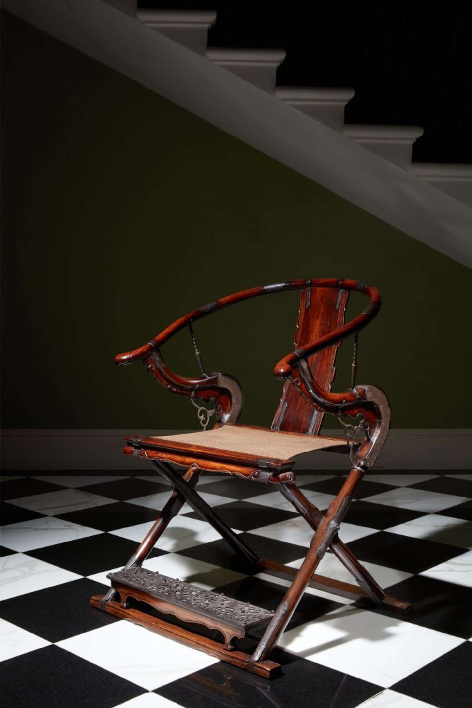 A decorative wooden chair with curved features and decorative foot rest attached on a black and white chequered floor