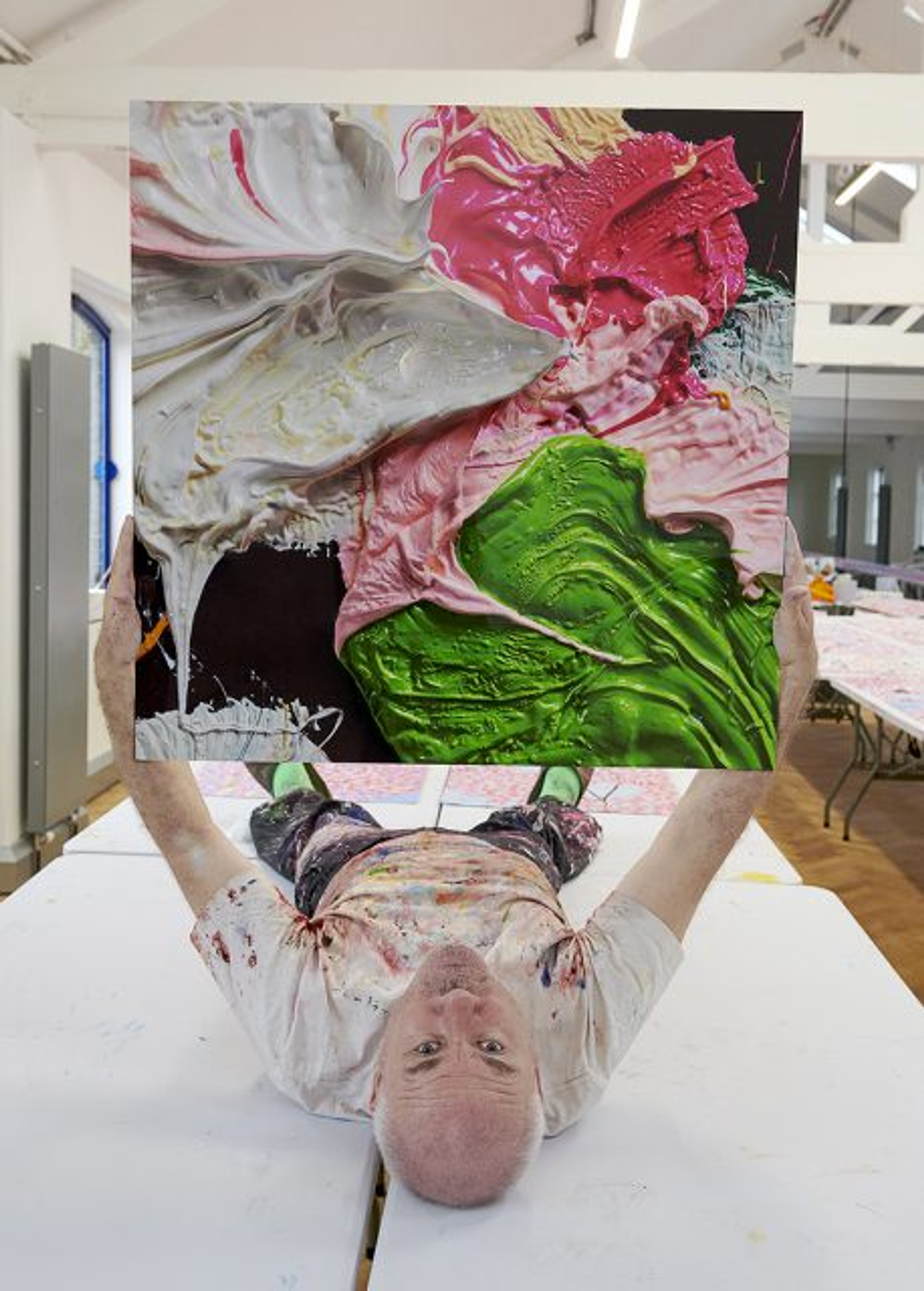 An image of the artist Damien Hirst, lying down and looking up at the viewer, holding one of his prints from the Fruitful And Forever series.