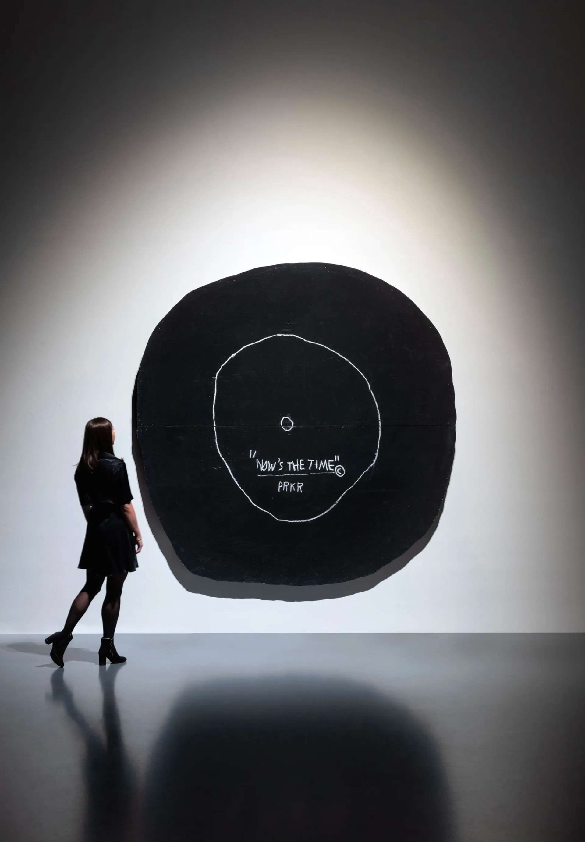 A woman standing beside and gazing at a large black rounded canvas resembling a record player. The artwork displays the phrase ”NOW'S THE TIME ©"