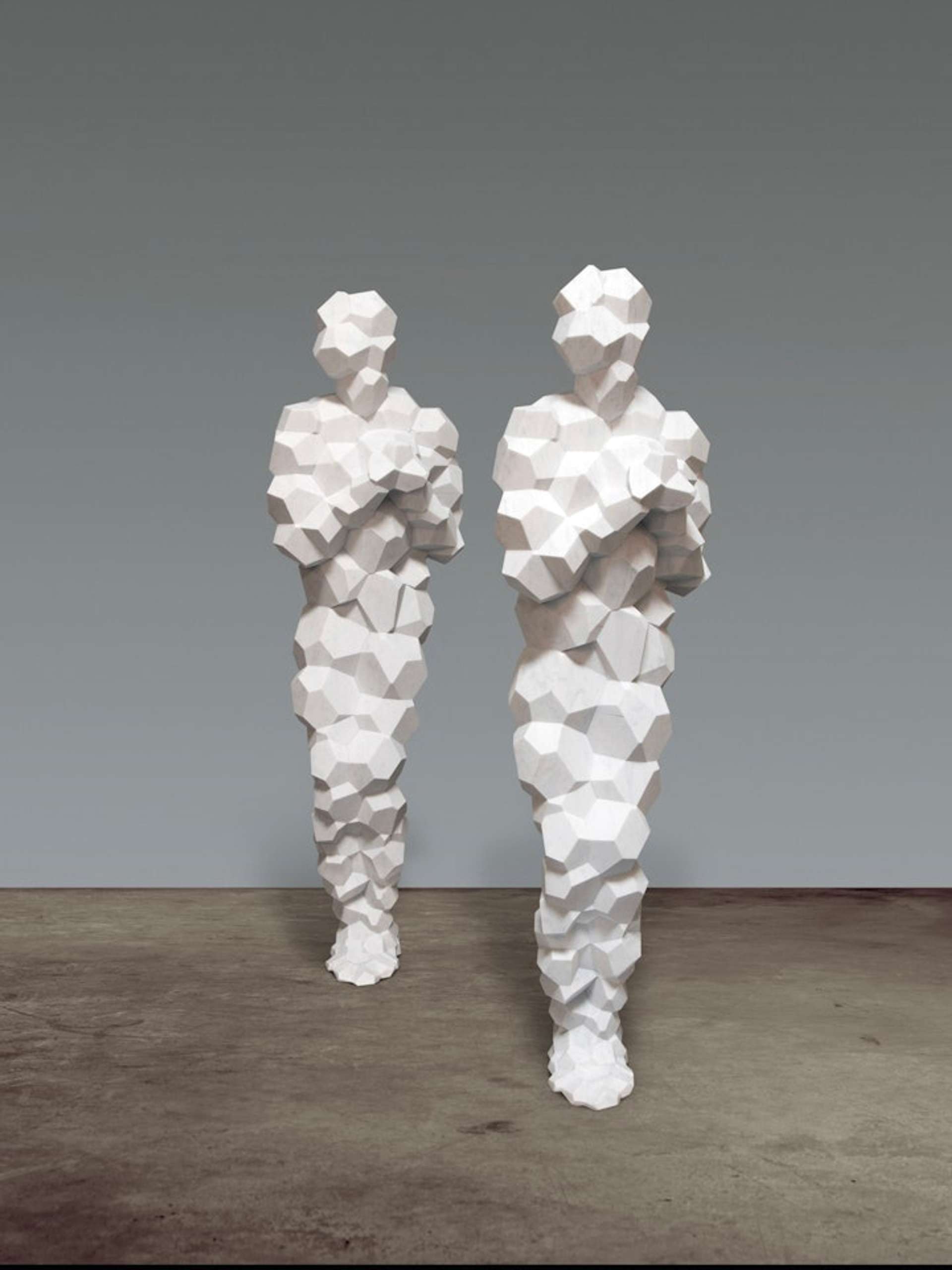 Two freestanding life-sized marble sculptures are captured in an unoccupied gallery, positioned on a concrete floor. The sculptures, crafted from marble, aim to portray the essence of the human form without any distinctive features. The marble material lends a textured surface to the figures, which are arranged in a staggered manner beside each other.