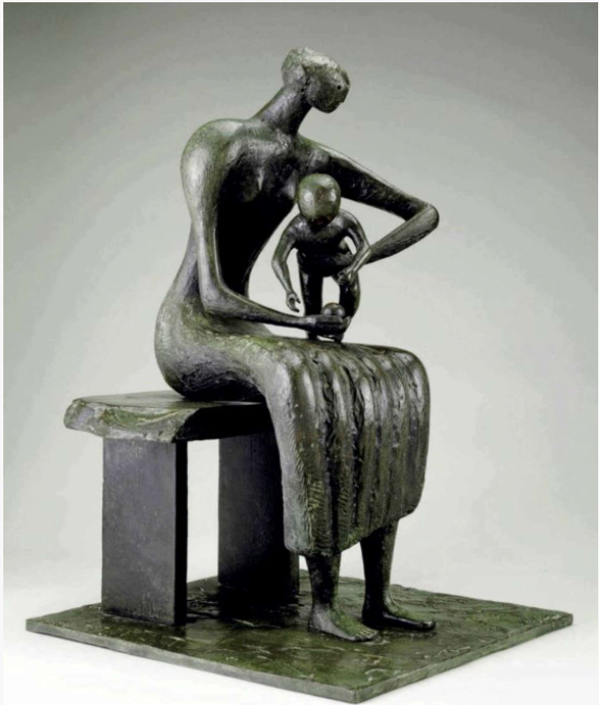 A bronze sculpture by Henry Moore depicting a seated woman cradling a standing baby on her lap. The woman supports the baby's torso with one arm while holding an apple in her other hand, which rests on her thigh and attracts the child's gaze. The sculpture showcases the woman seated on a bench and is presented on a thin square plinth.
