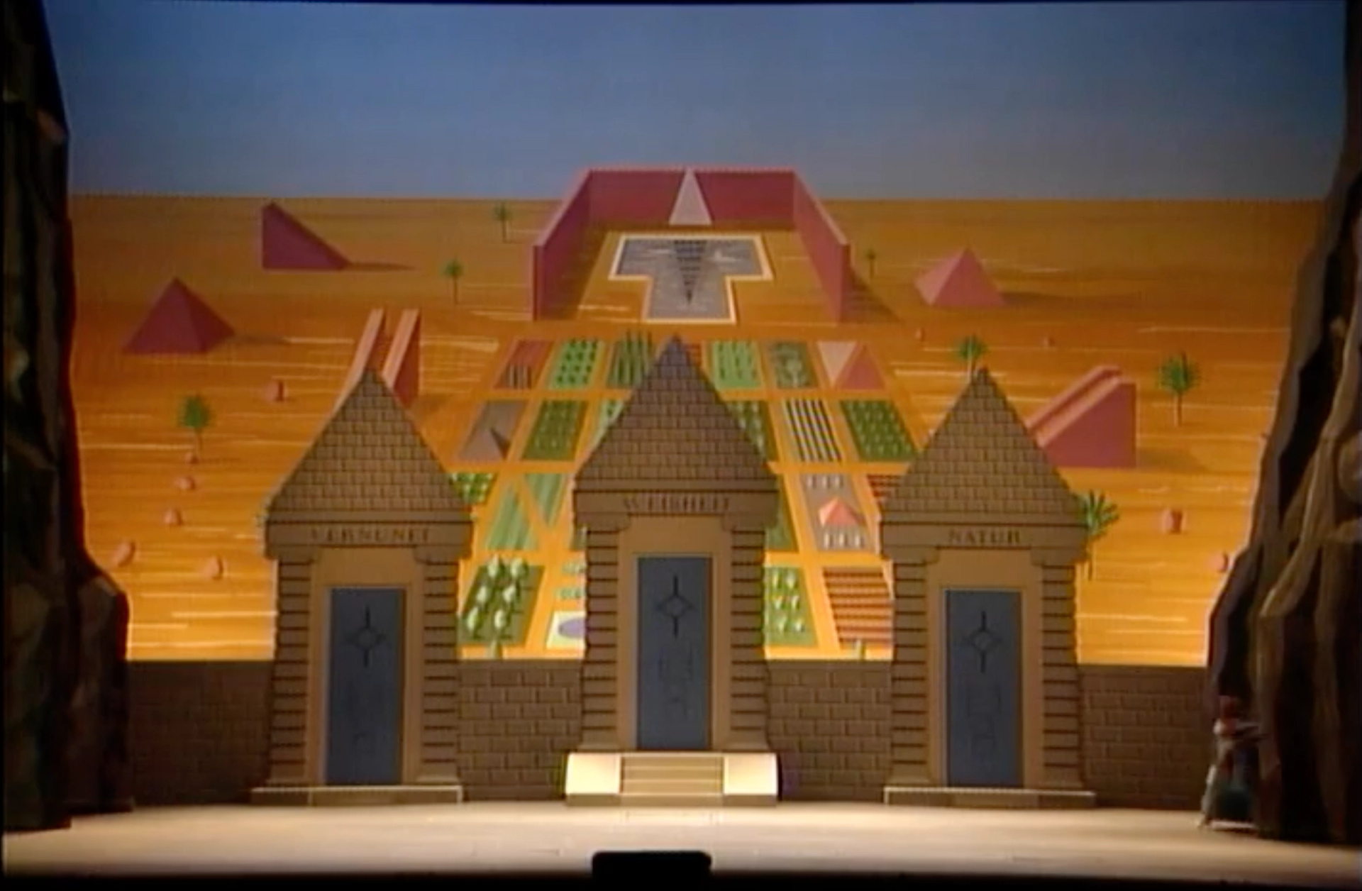 An image of a set designed by David Hockney for the Magic Flute. It features three gates on a wall leading to a landscaped garden.