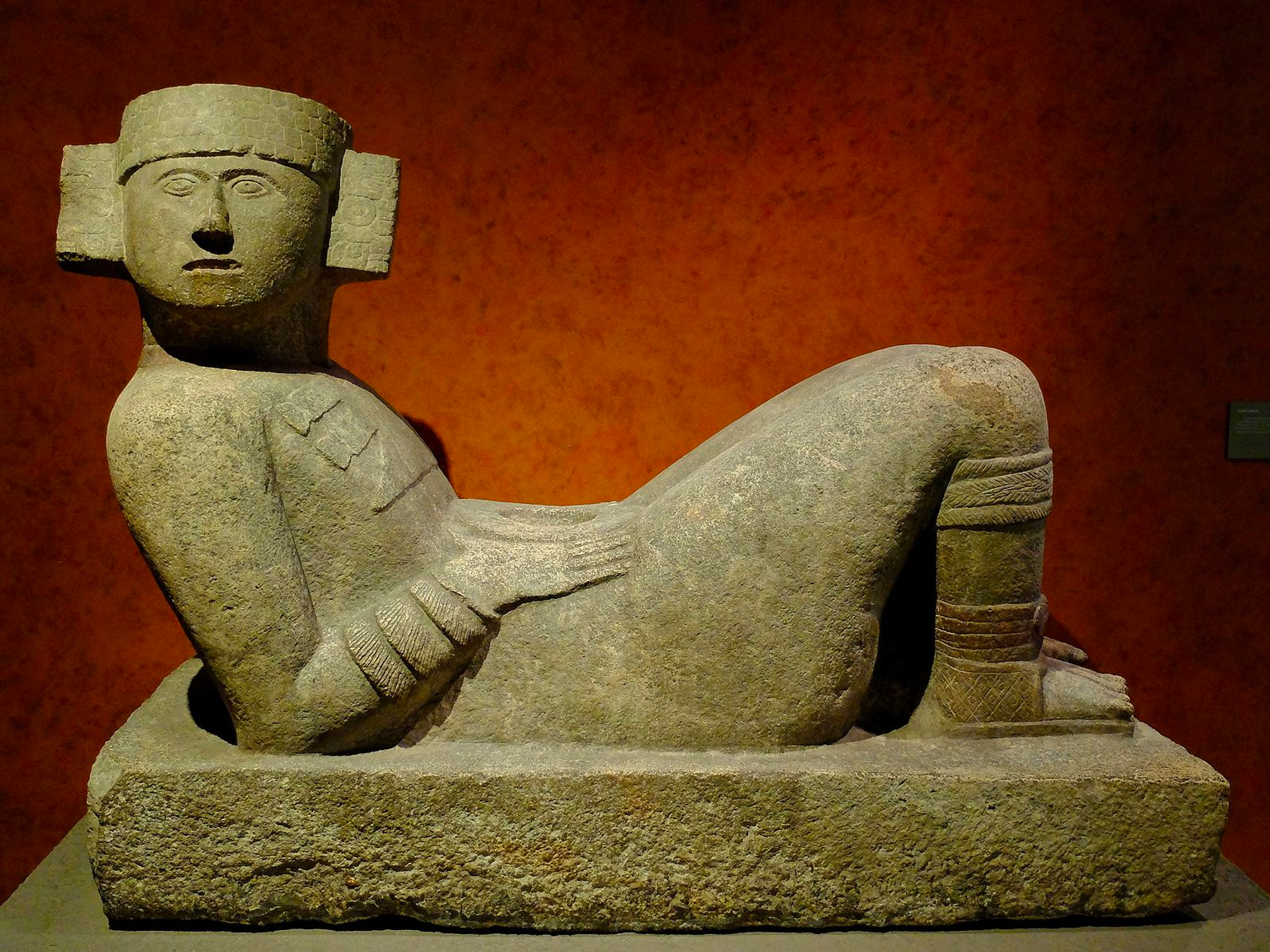 Abstract sculpture of a reclining figure with knees propped up, resting on forearms. The sculpture's head faces the viewer, adorned with a headdress. Imprints of decorative sandals are visible, extending up to the knees. The figure's hands rest on its lap.