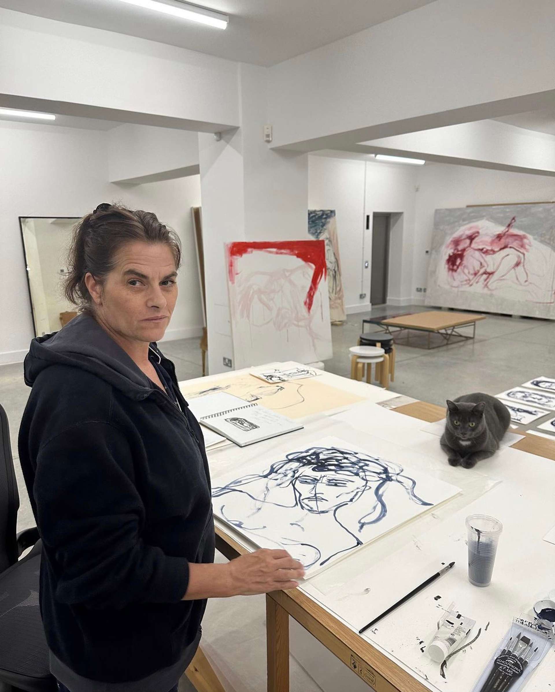 An image of the artist Tracey Emin in her studio, drawing the works that were engraved on the bronze doors. The artist is looking into the camera as a cat lies in the background.