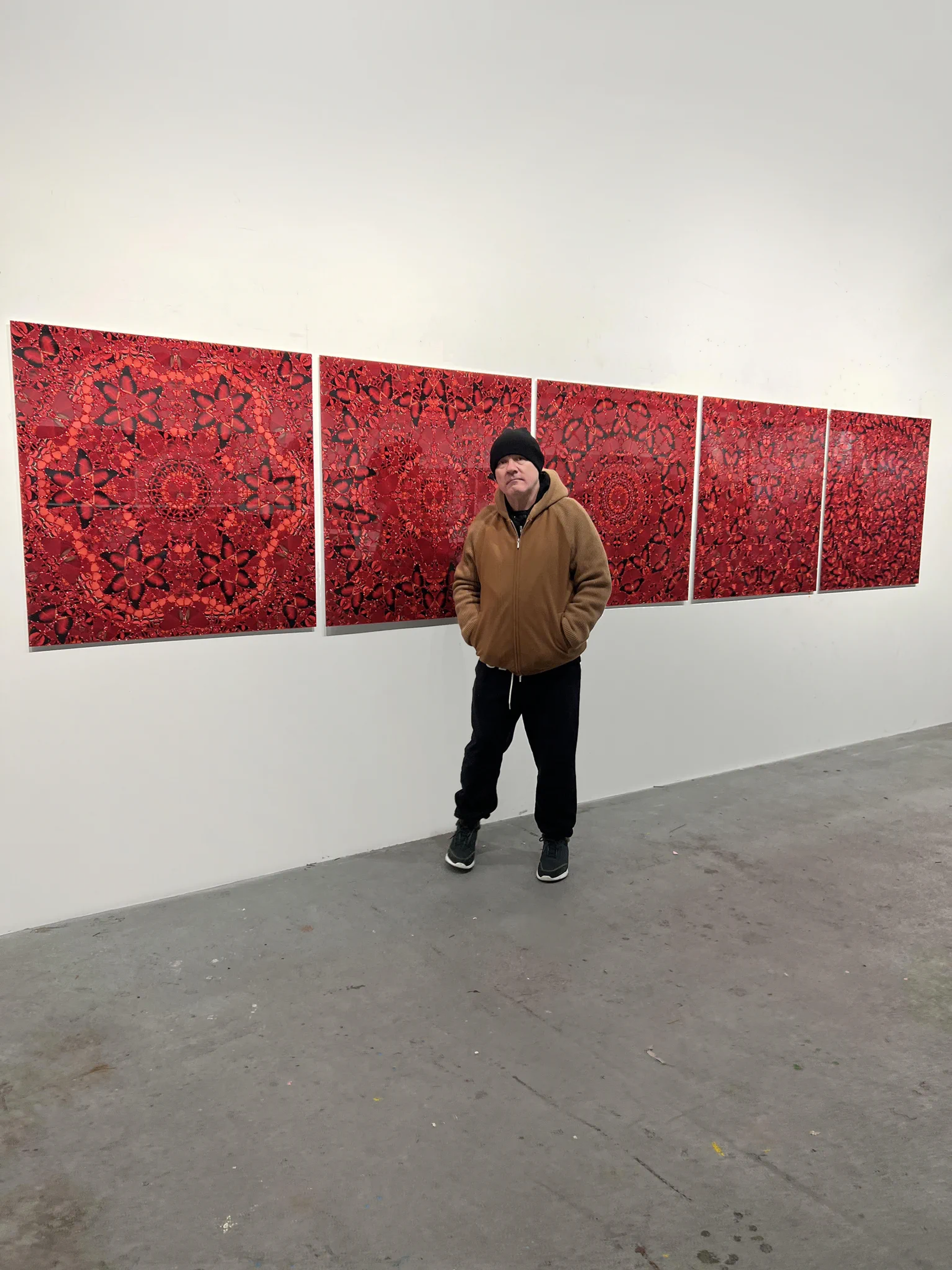 An image of the artist Damien Hirst standing in front of his Empress series. He is shown wearing a beige hoodie and a black beanie and jeans, with his hands in his pockets.