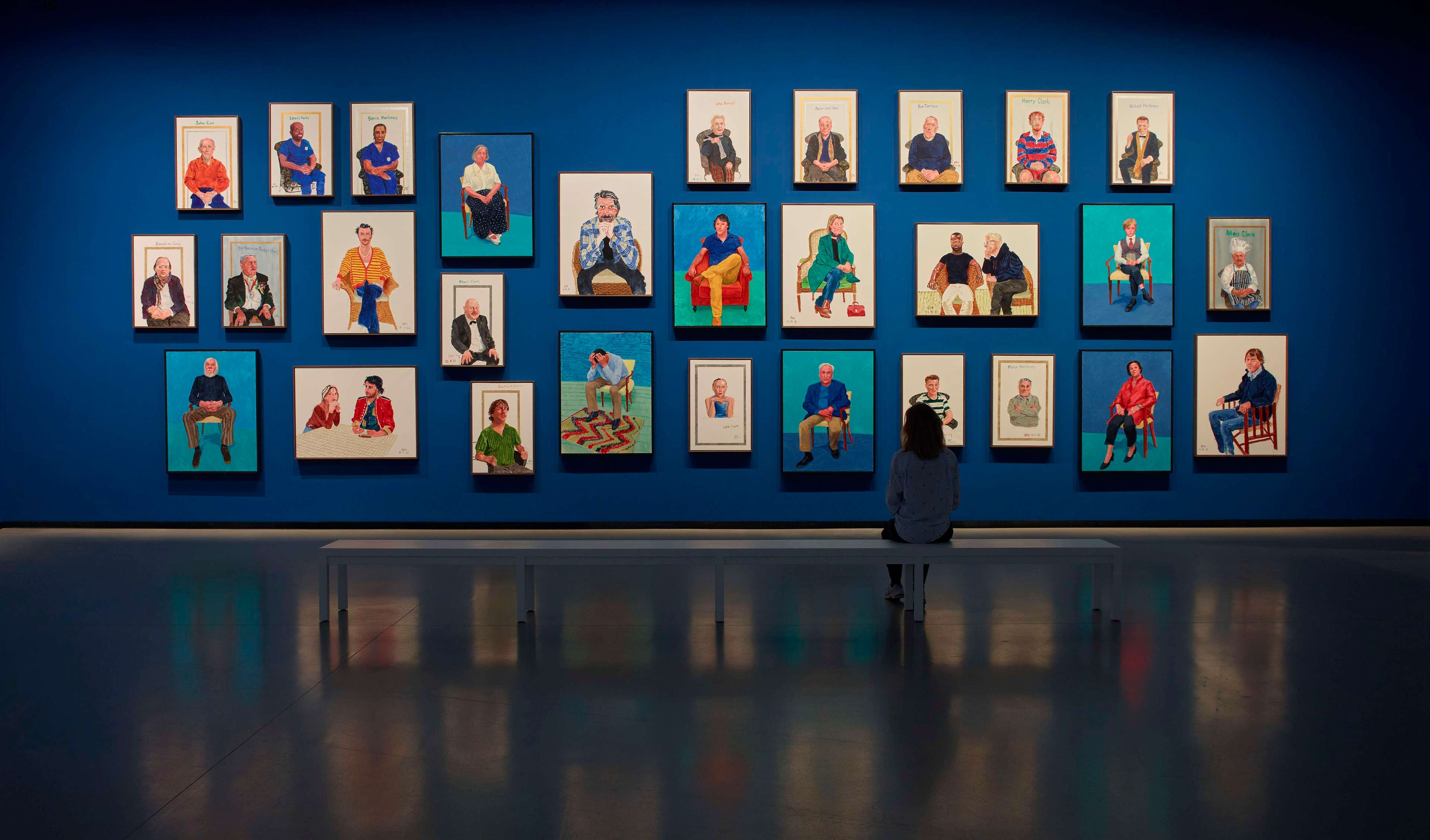Visitor seated on a bench facing a salon-style wall of David Hockney portraits against a vivid blue backdrop at Fondation Louis Vuitton, Paris.