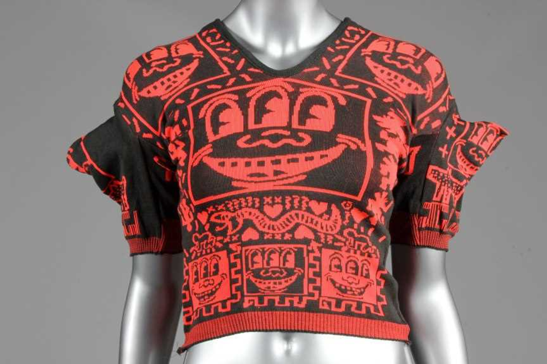 An image of a mannequin wearing a black and red knitted t-shirt, printed with Keith Haring's three-eyed smiley face motif.