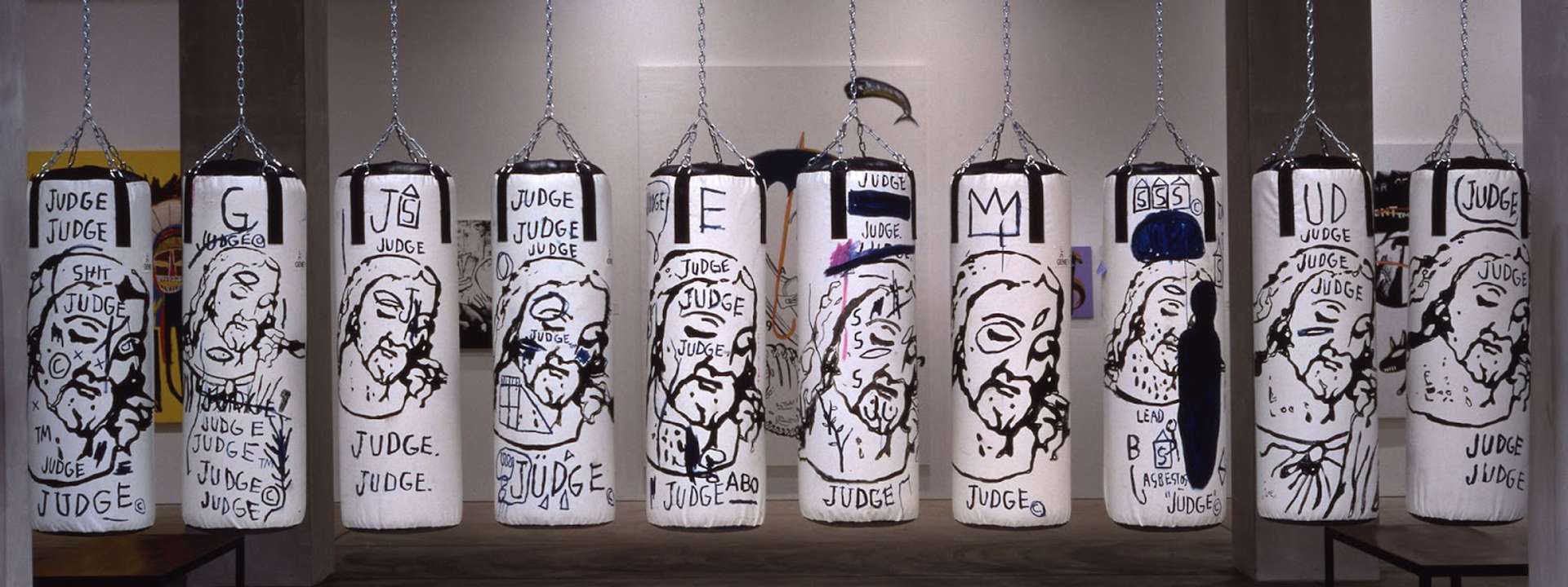 The artwork Ten Punching Bags by artists Jean-Michel Basquiat and Andy Warhol, showing ten white punching bags hanging from the ceiling. In each, a Renaissance-like depiction of Jesus is painted, and graffitied over by Basquiat with the word “Judge”.