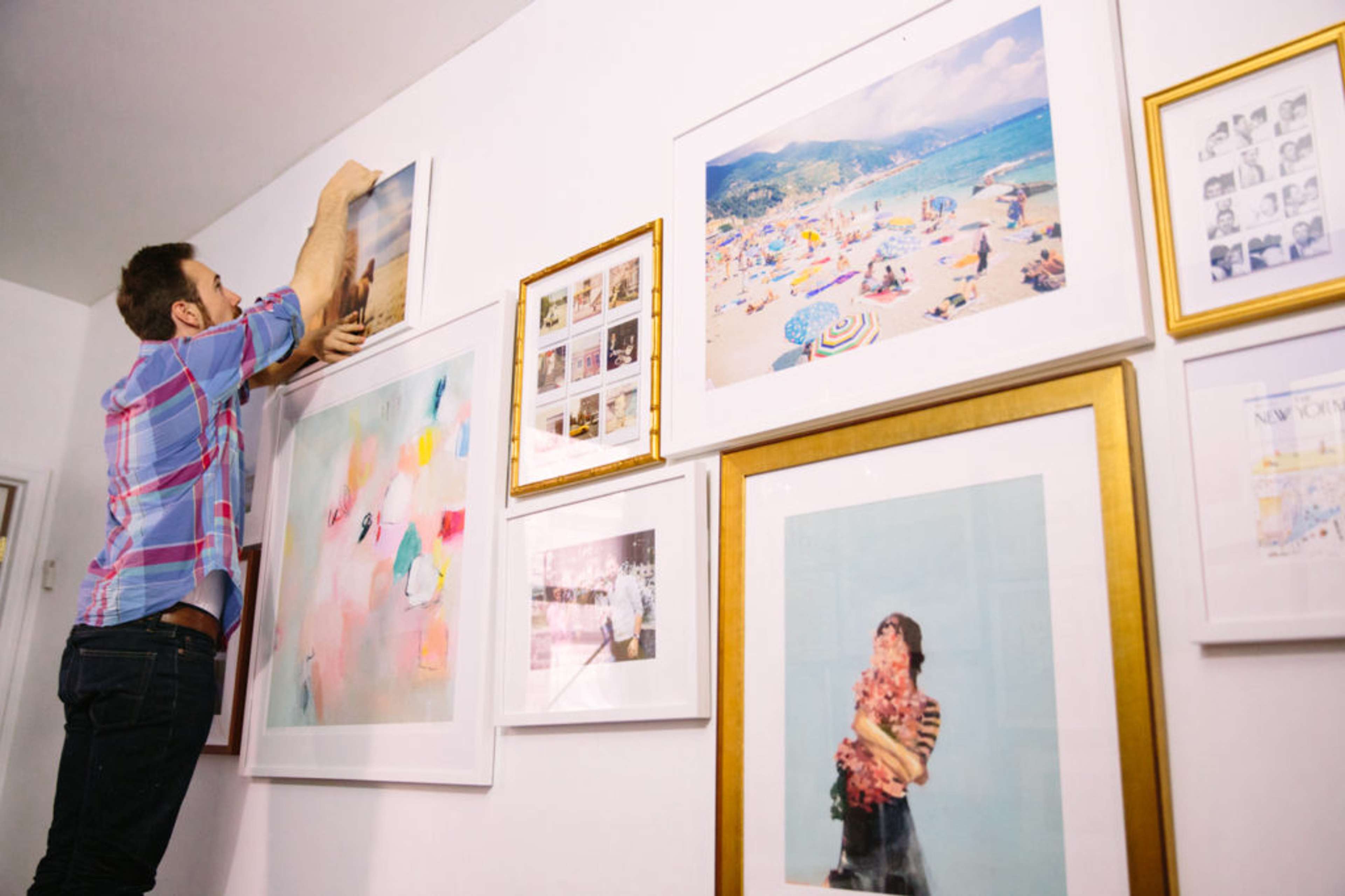 A man on a ladder putting artwork on a white wall that already has multiple artworks mounted on it 