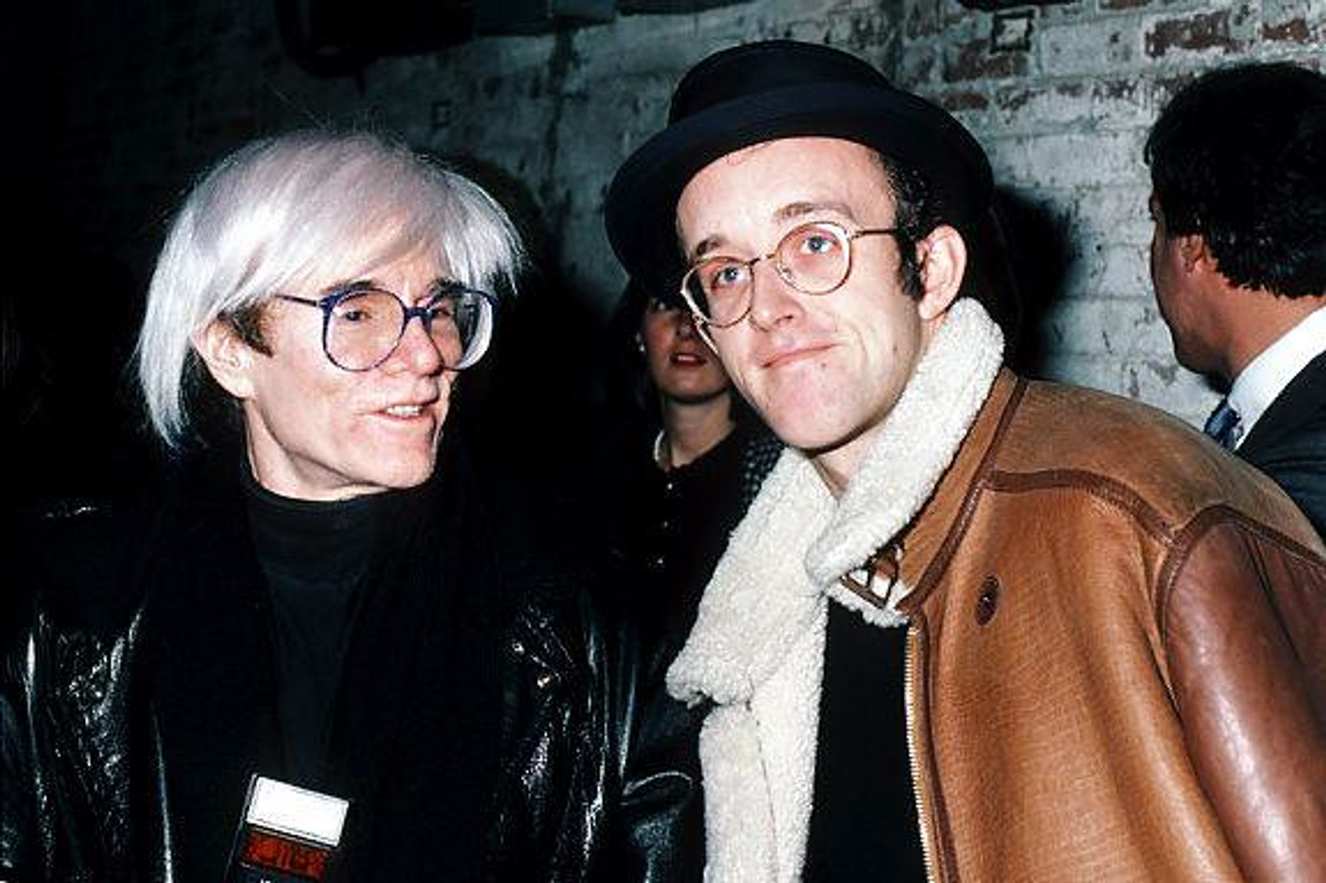 A photograph of the artists Andy Warhol and Keith Haring together. Haring smiles at the camera while Warhol talks to him.
