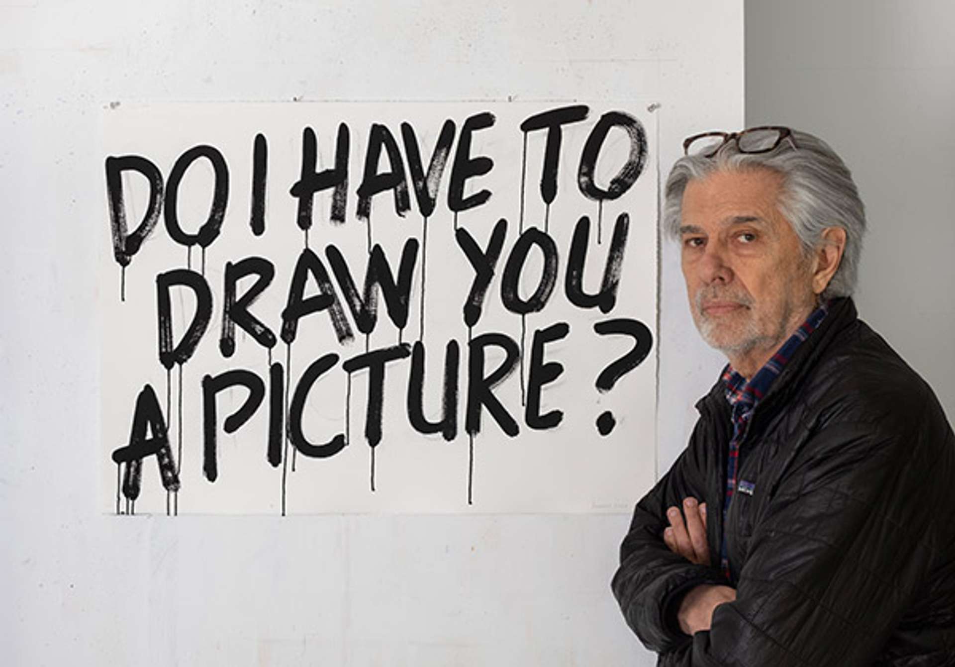 A photograph of Mel Bochner standing against a white wall, crossing his arms and turning his head towards the viewer. The wall is adorned with the spray-painted phrase "DO I HAVE TO DRAW YOU A PICTURE?" in bold black letters.