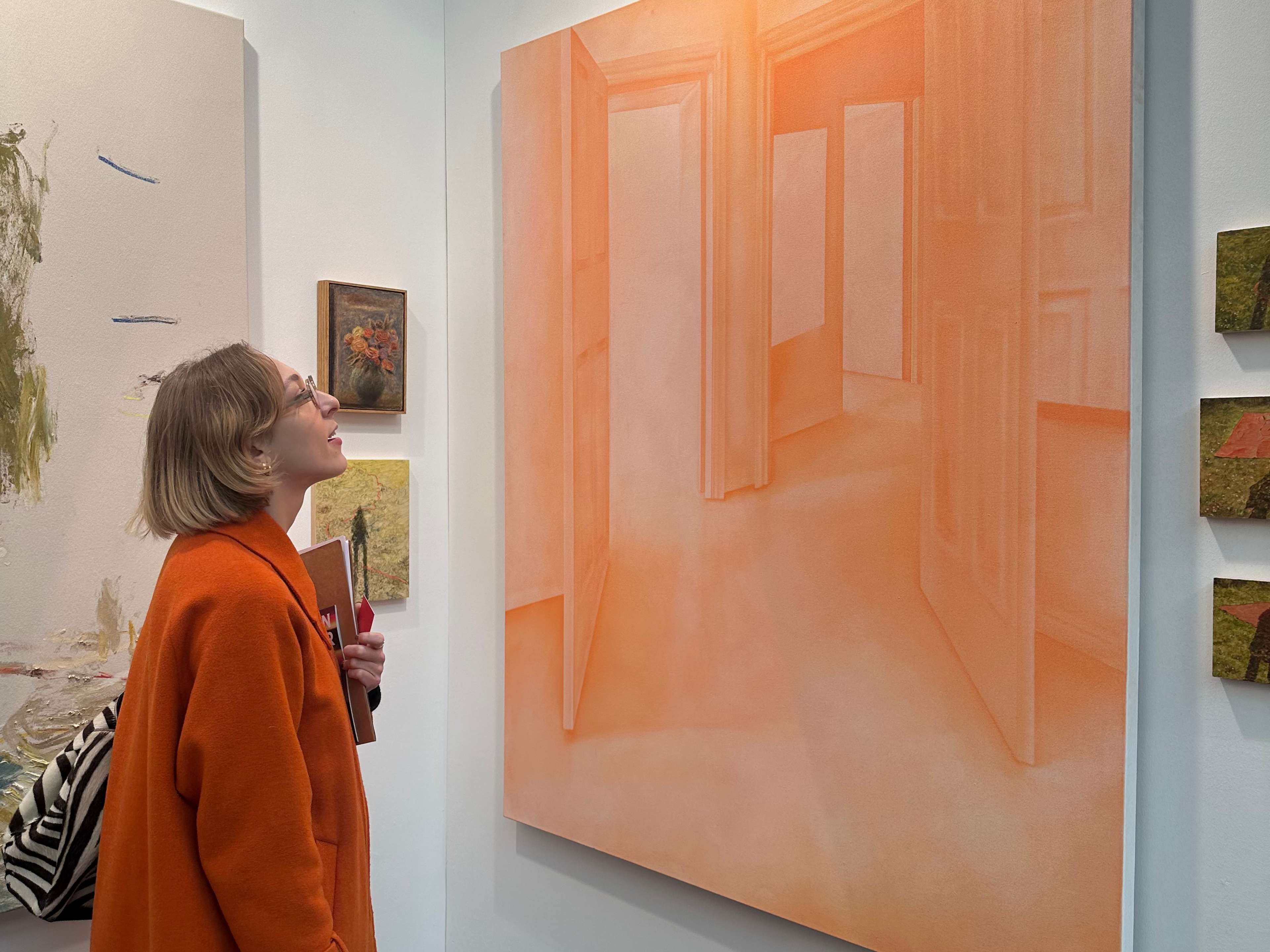 A young woman wearing a long, orange coat gazes up at a large canvas hanging on a wall