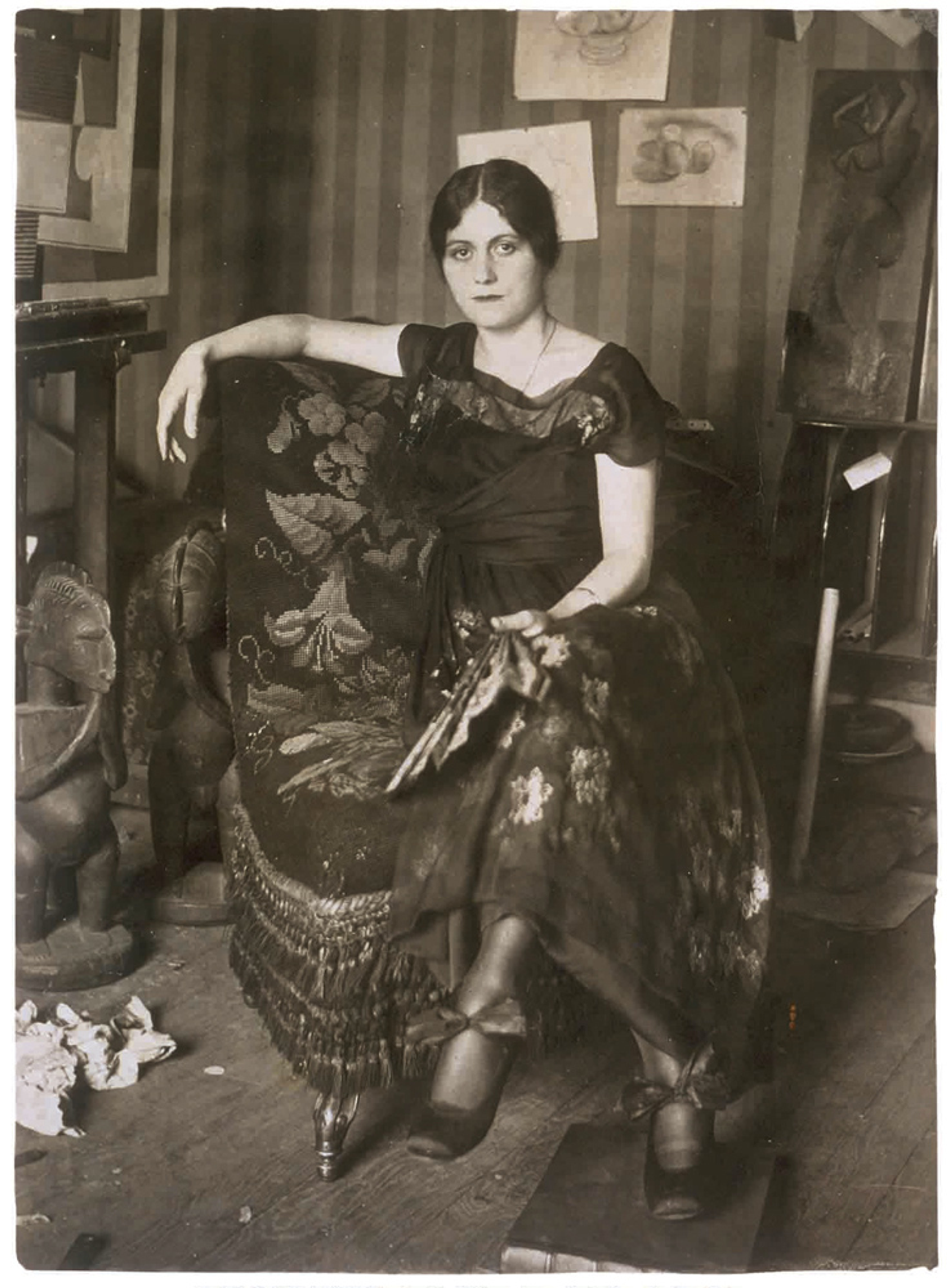 A black-and-white photograph of Olga Khokhlova in Picasso's Montrouge studio. She is shown sitting with her legs crossed while holding a fan.