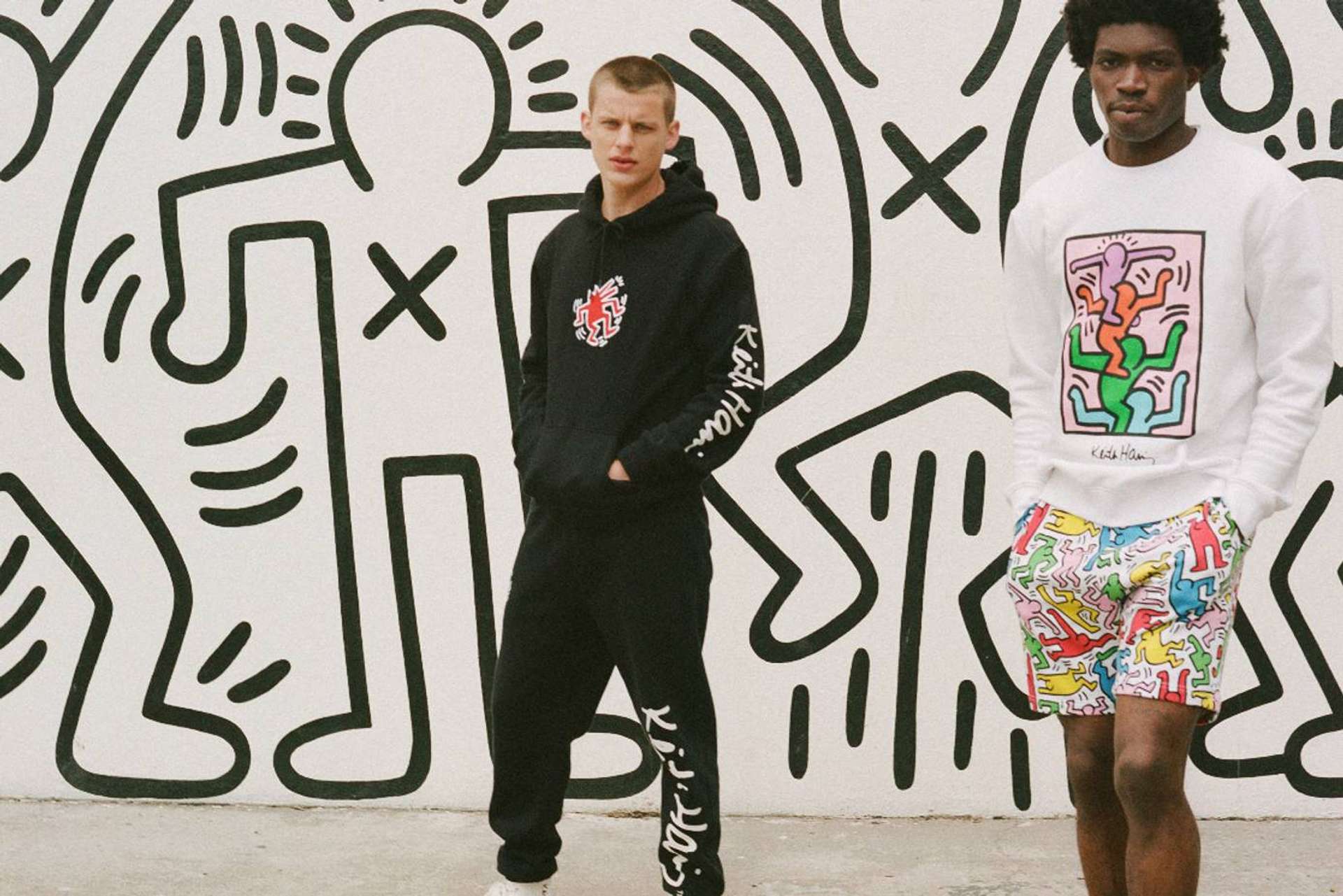 An image of two models wearing apparel from H&M’s collaboration with Keith Haring, standing against a background featuring one of the artist’s iconic murals.