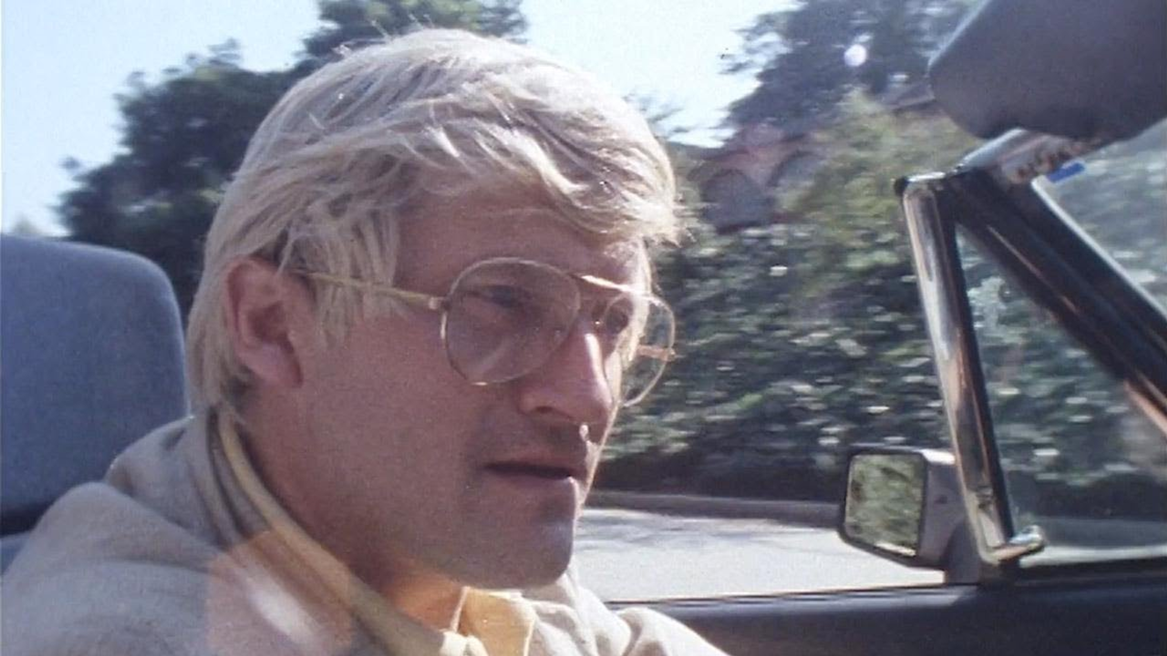 This screenshot shows the artist David Hockney, still young, driving a convertible car.