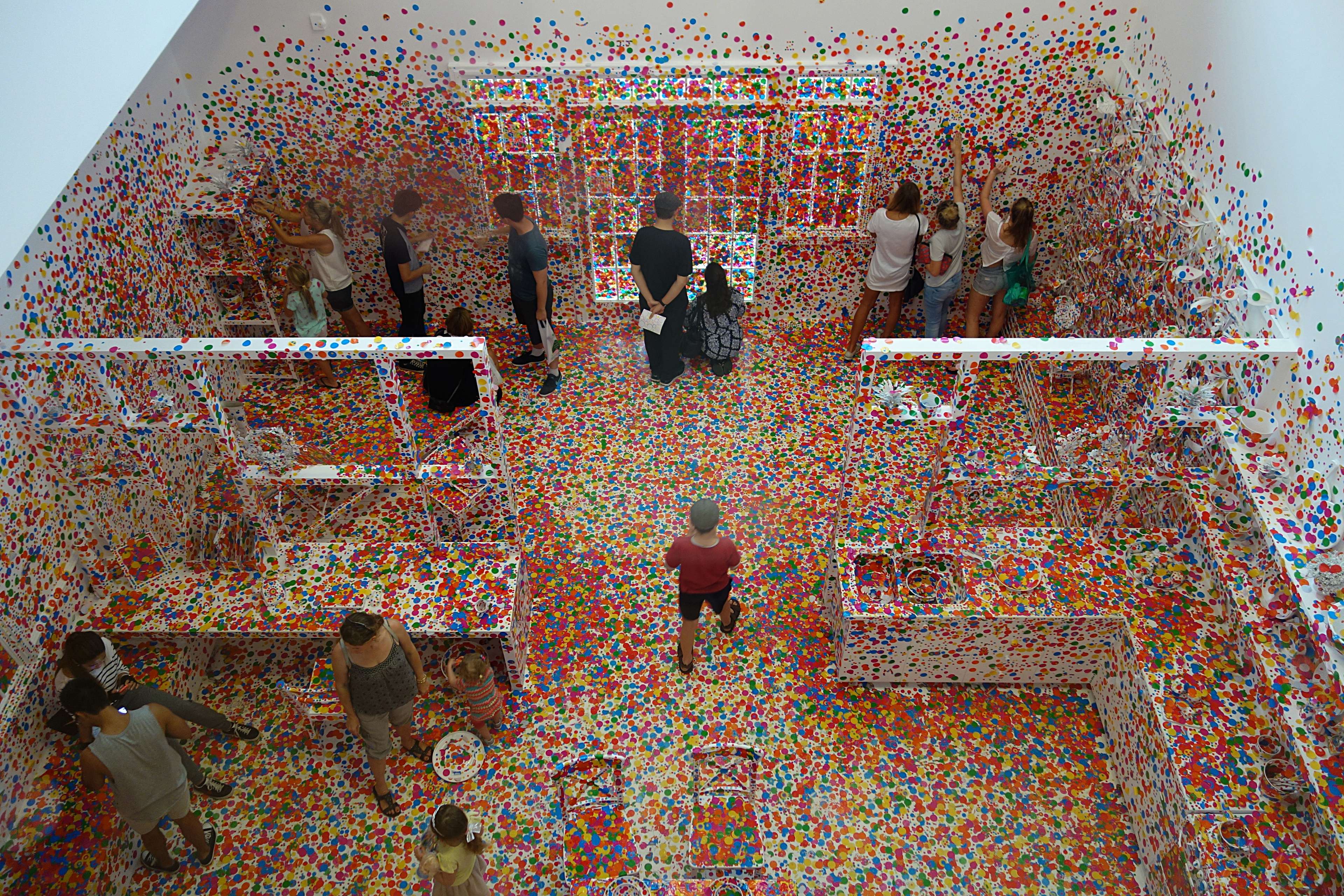 A photograph shot from a birds eye view, depicting a white room with white furniture largely covered in colourful stickered dots. People are seen walking around and attaching the dots to the walls and furniture.