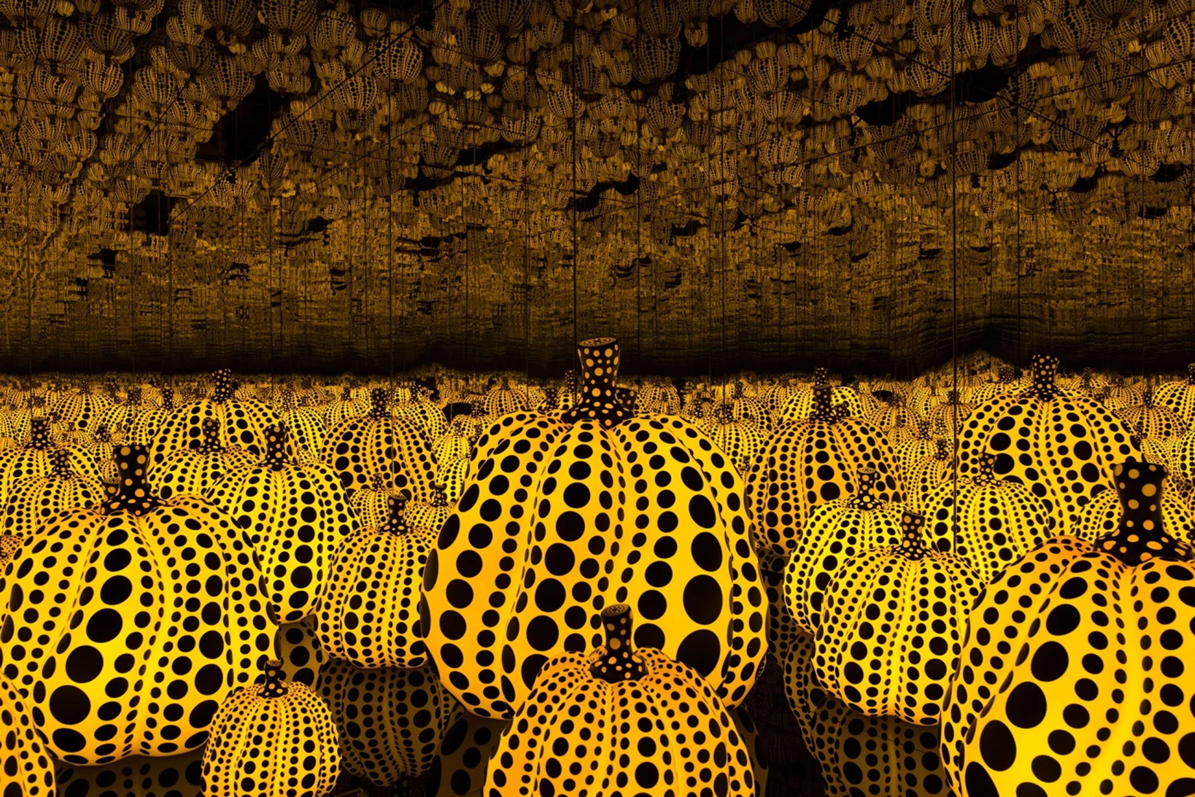 A photograph depicting a completely mirrored room, endlessly reflecting images of a variety of differently sized glowing yellow pumpkin sculptures, covered in black dots. 