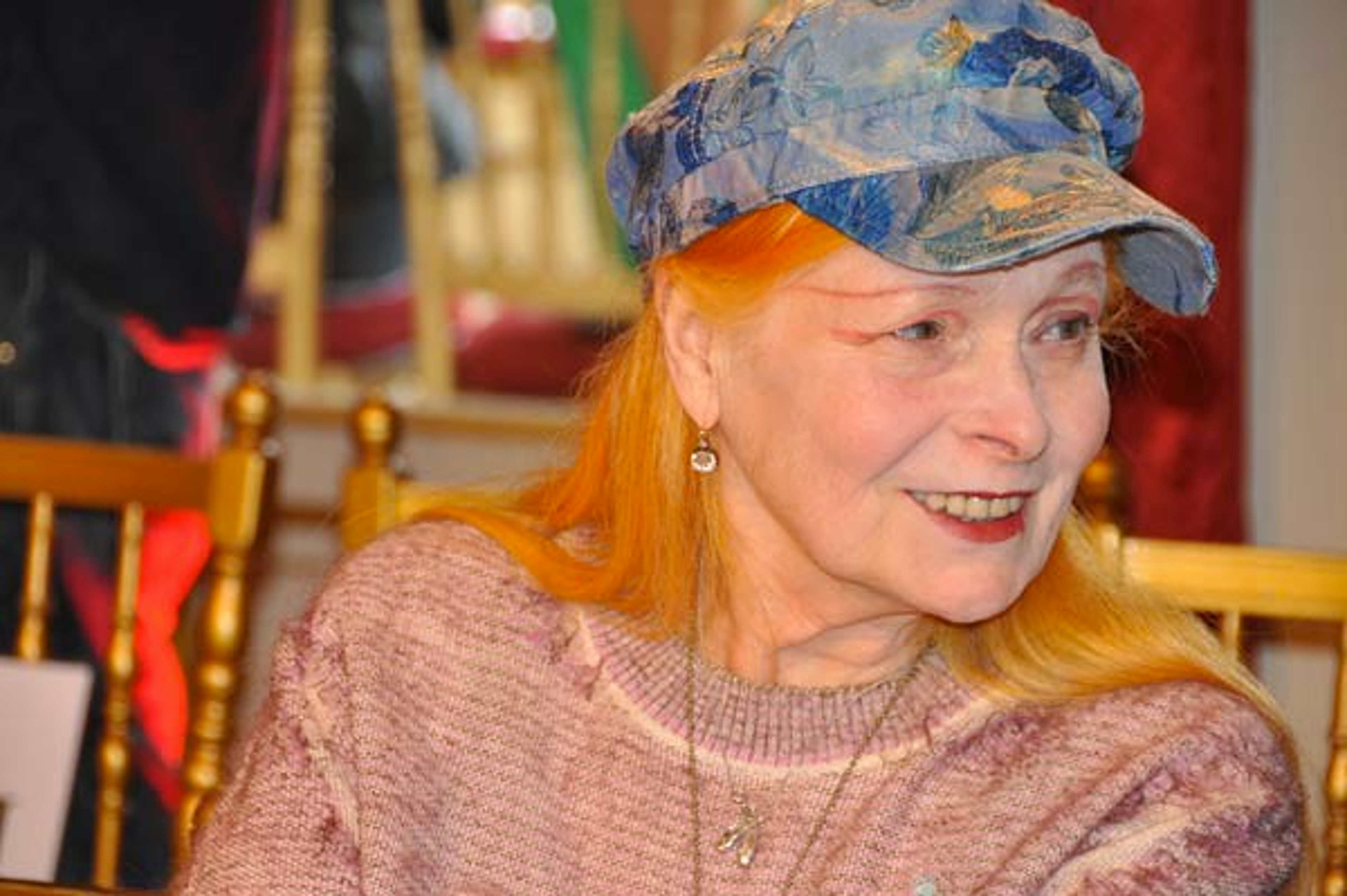 An image of the designer Vivienne Westwood, with her signature orange hair. She is wearing a blue jeans hat, and a distressed pink jumper. She is smiling at someone off-camera.
