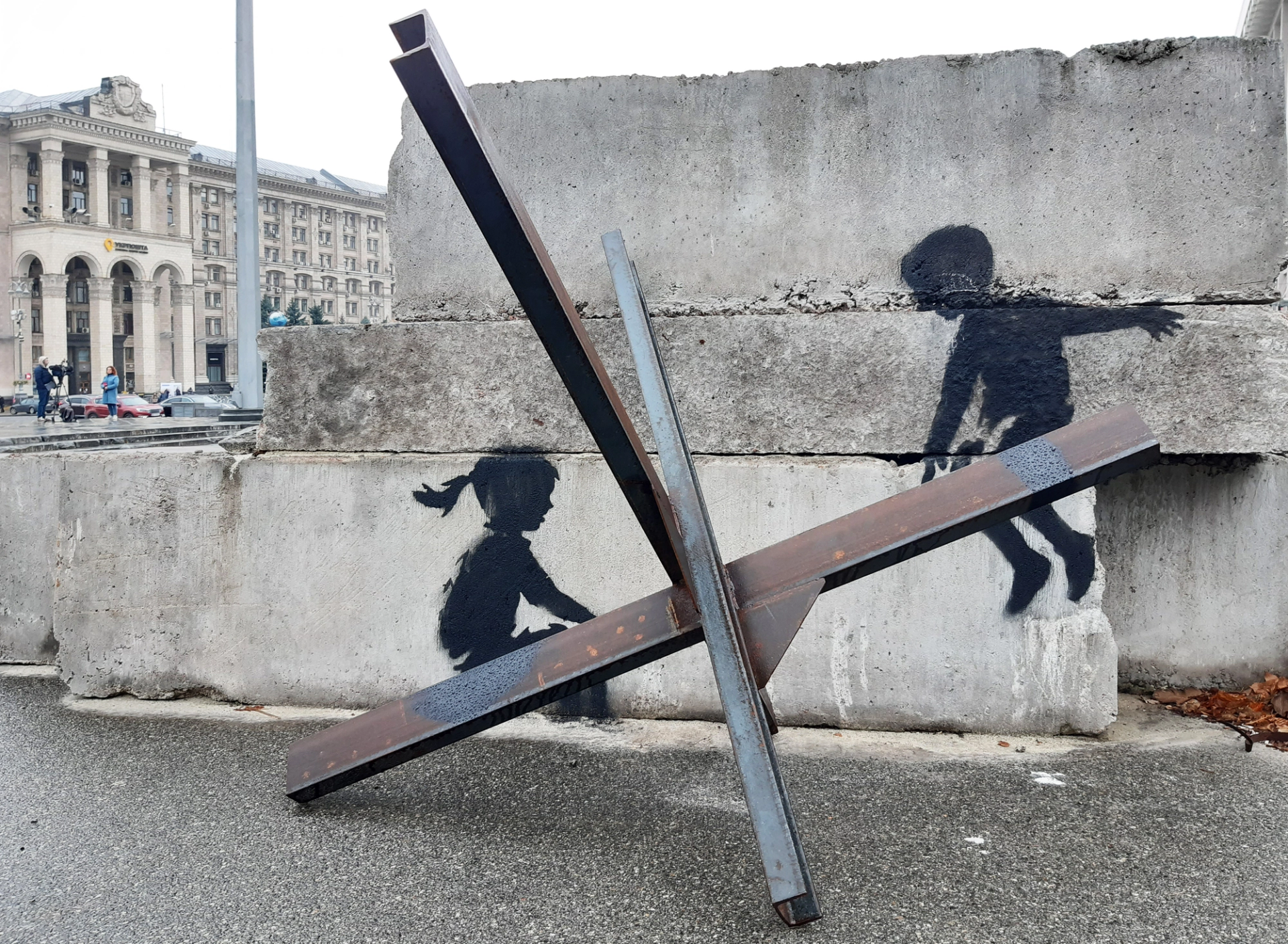 An image of a Banksy mural in Independence Square, Ukraine. It shows the silhouette of two young children, a boy and a girl, playing on a seesaw made out of shrapnel.