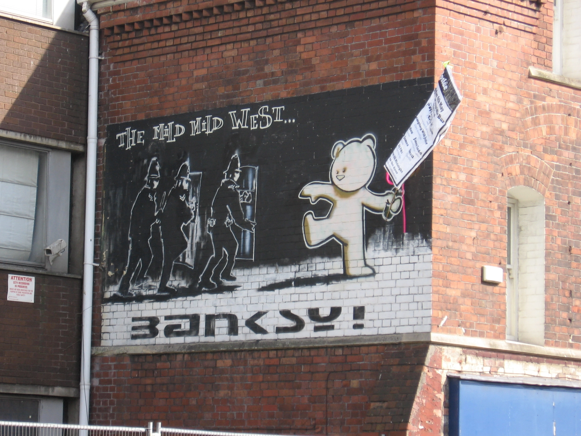 An image of the mural Mild Mild West by Banksy. It shows a soft, plush-like teddy bear throwing a firebomb on a group of police officers.