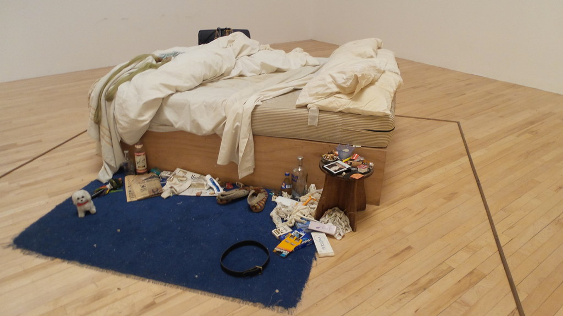 An image of Emin's bed, unmade and surrounded by debris including a bottle of vodka, such as condoms, underwear with menstrual blood stains, and functional objects, including a pair of slippers.