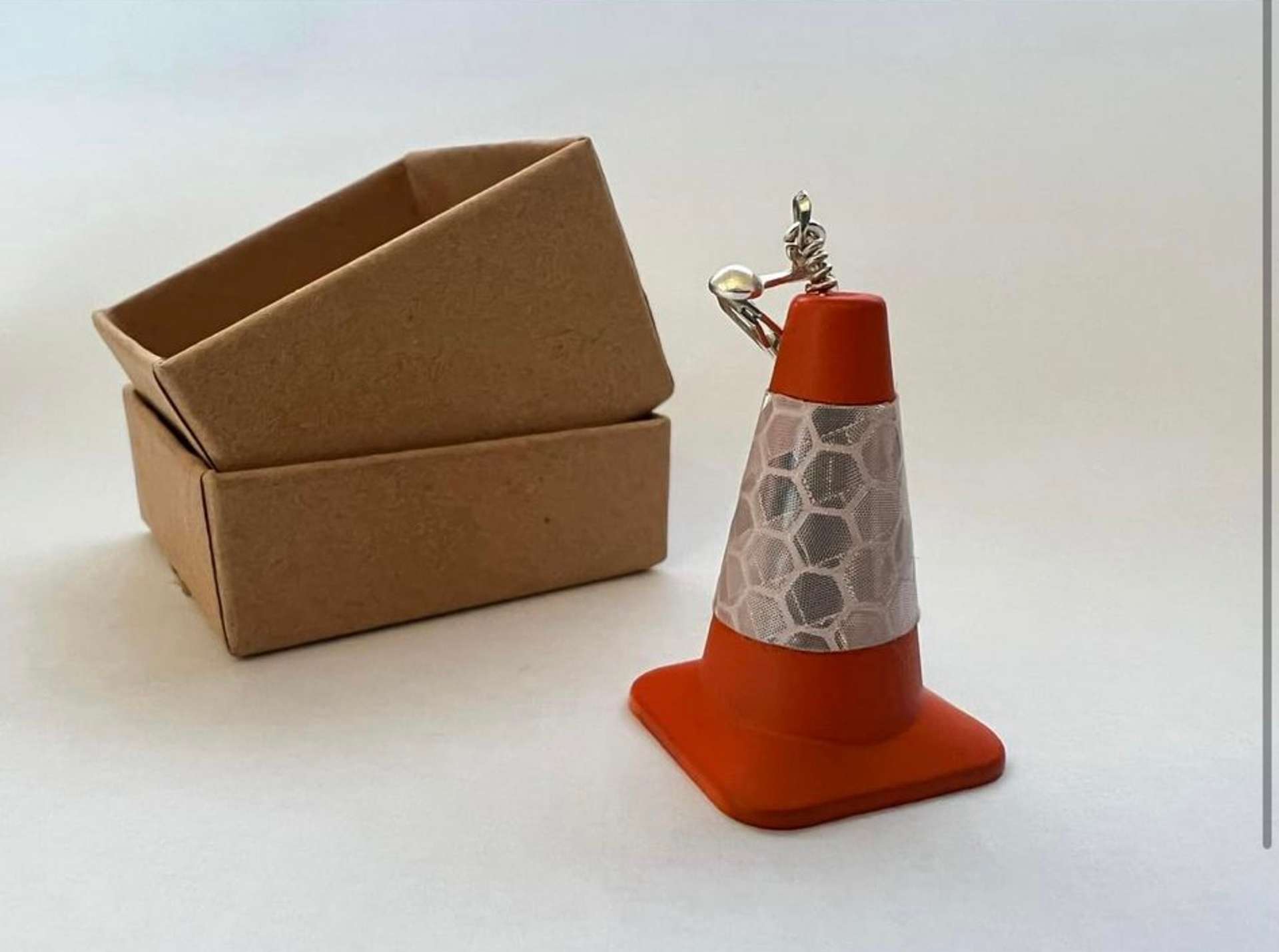 A photograph of a novelty earring in the shape of an orange traffic cone, next to a small box