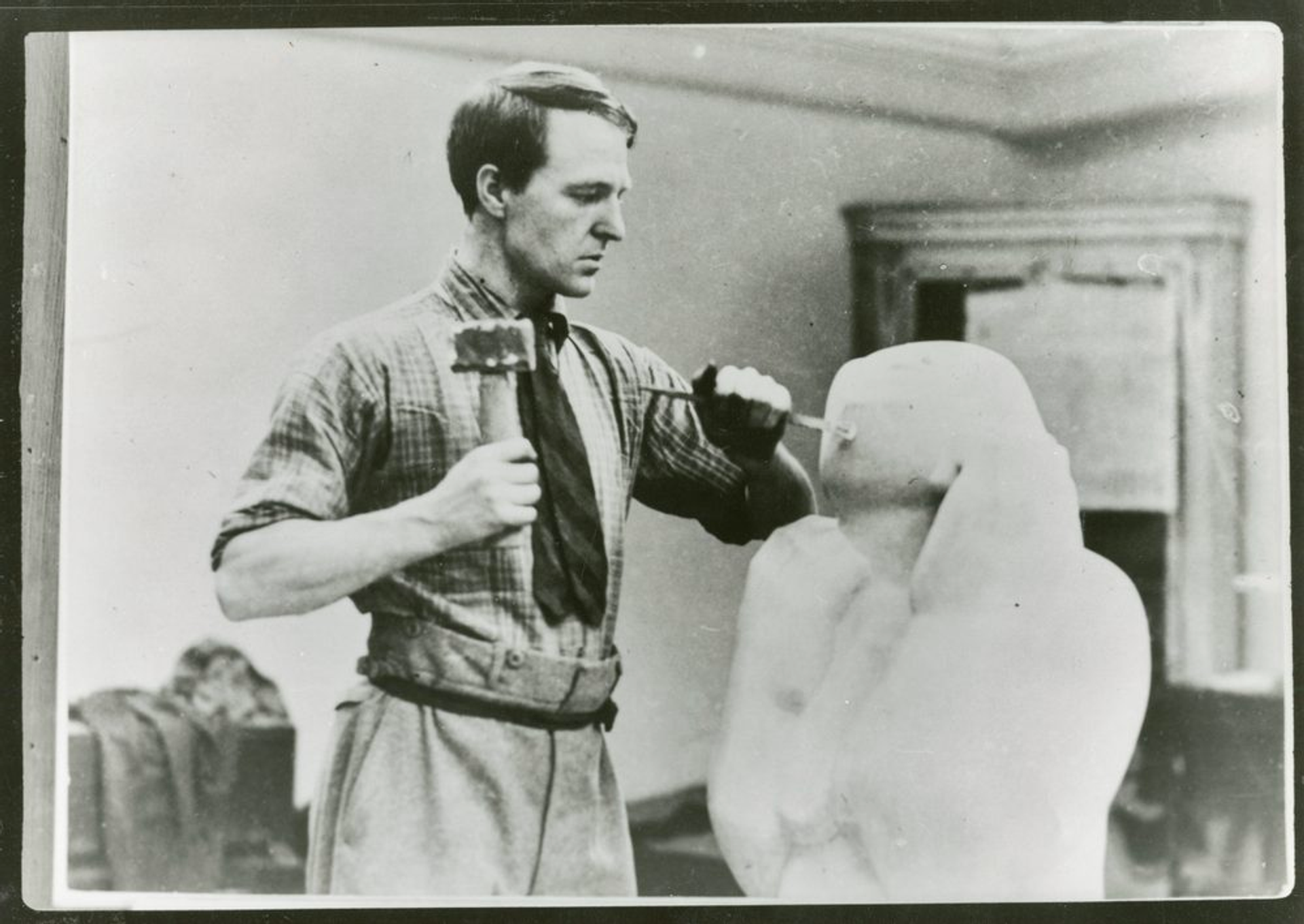  A black-and-white photograph capturing a young Henry Moore in his studio, actively engaged in sculpting. Moore is seen holding a hammer in one hand and a chisel in the other, seemingly working on the head of a sculpture that is still in progress.