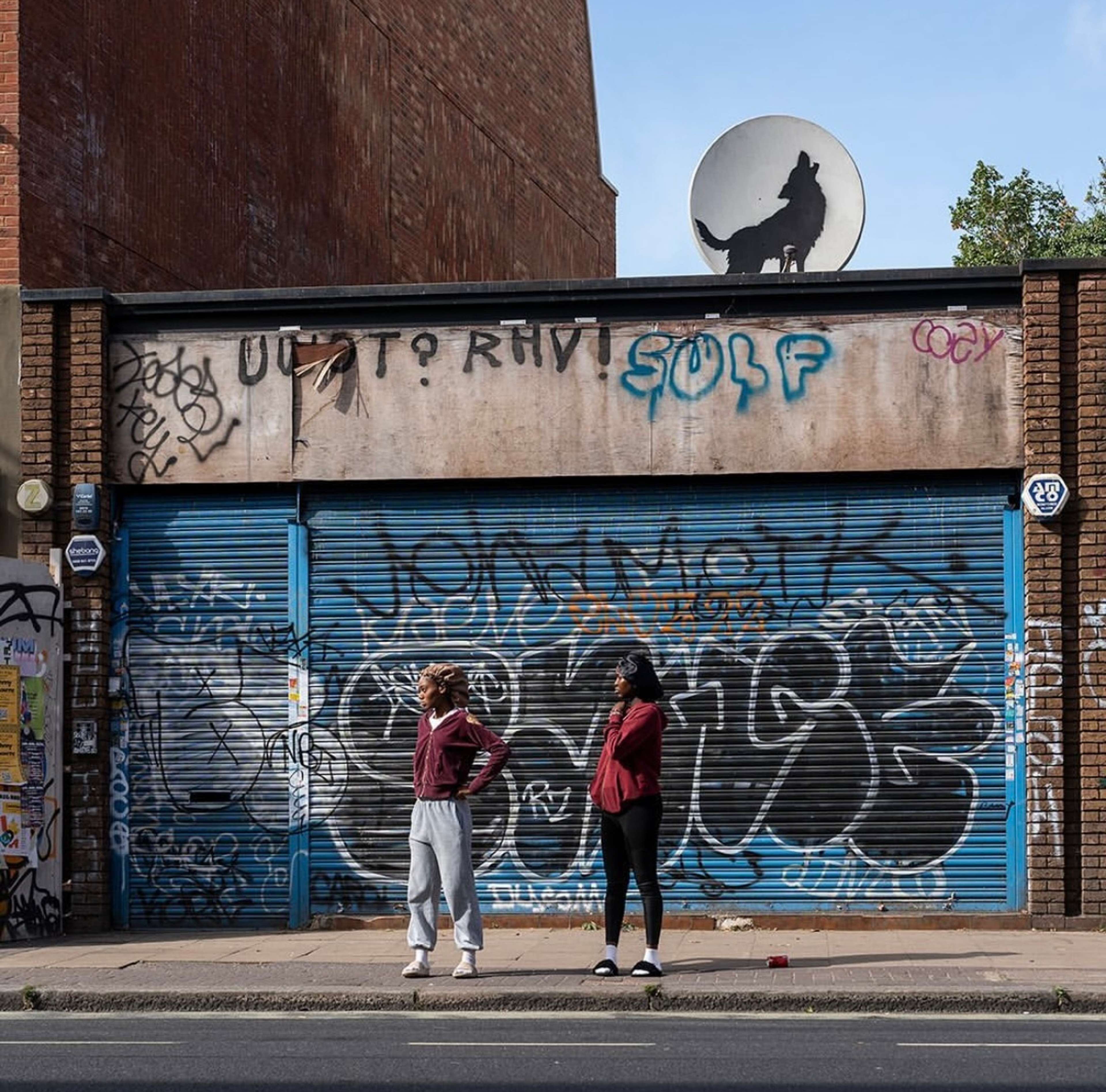 Banksy's lone wolf artwork spray painted on a satellite dish above a building covered in graffiti in Peckham, London. Two passers-by stand in front of the building wearing red hoodies.