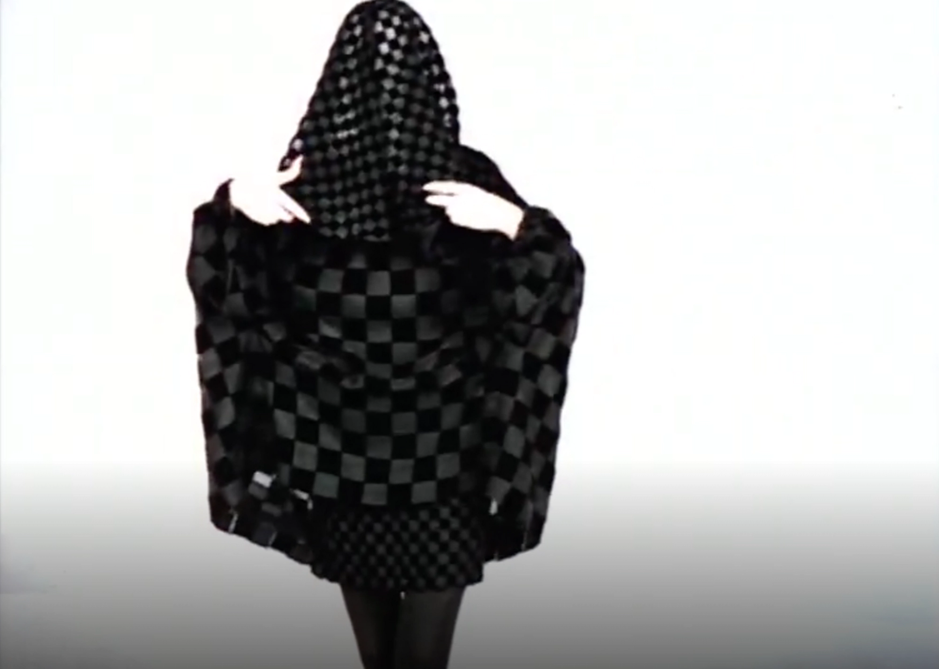 A screenshot of a veiled woman in black against a white background. Her entire garb is patterned with black squares of different sizes. 