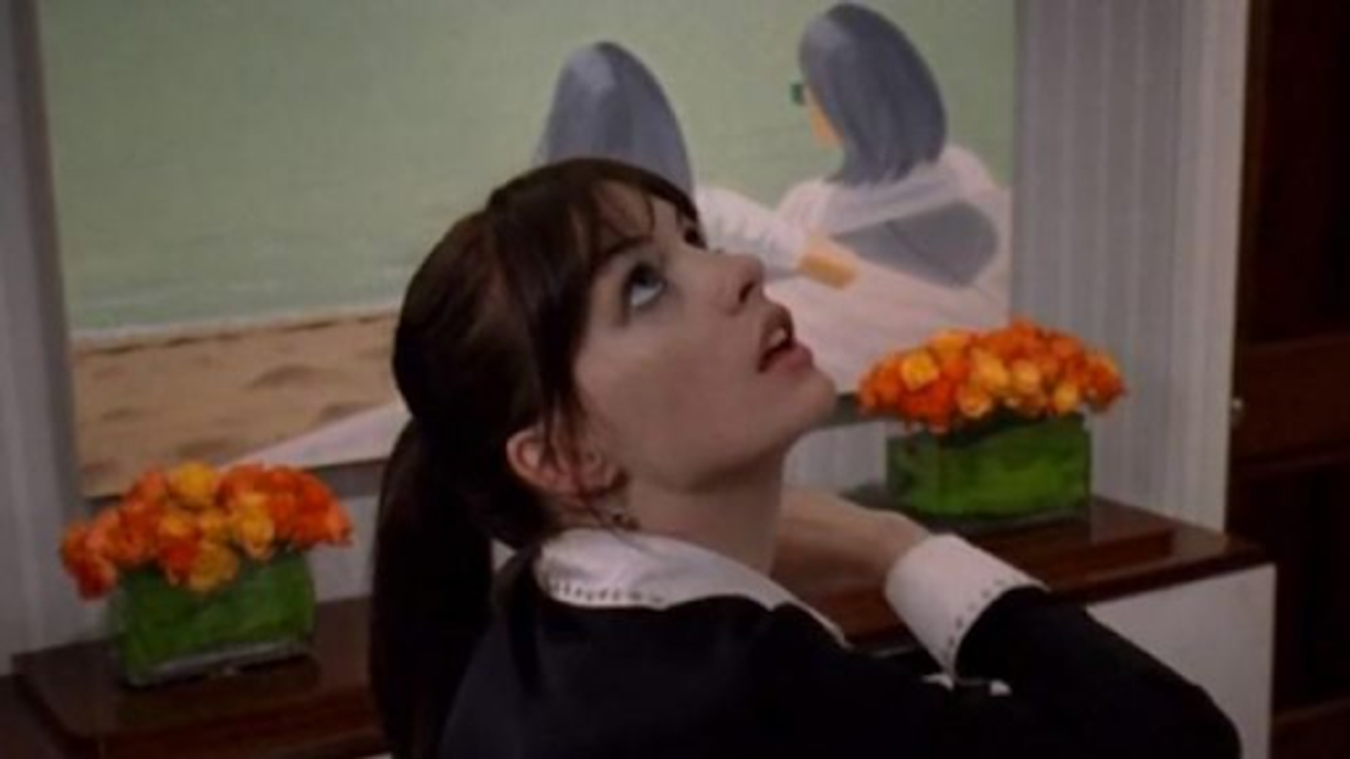 Photograph from the film The Devil Wears Prada. Woman looking up with a painting and floral arrangements on a table behind her. 