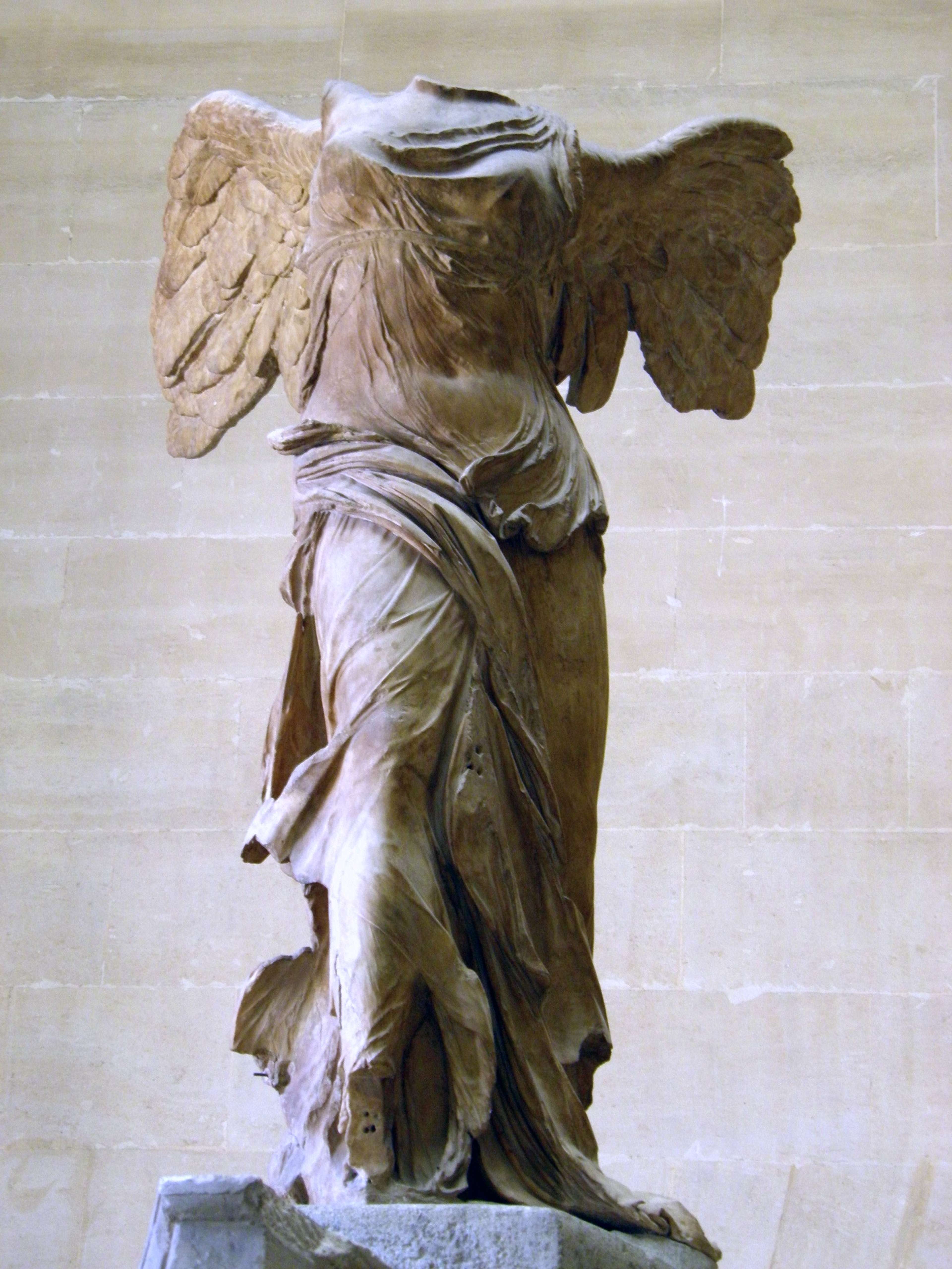 A headless figure with outstretched wings stands poised on a stone base, its flowing drapery carved to suggest wind-swept motion.