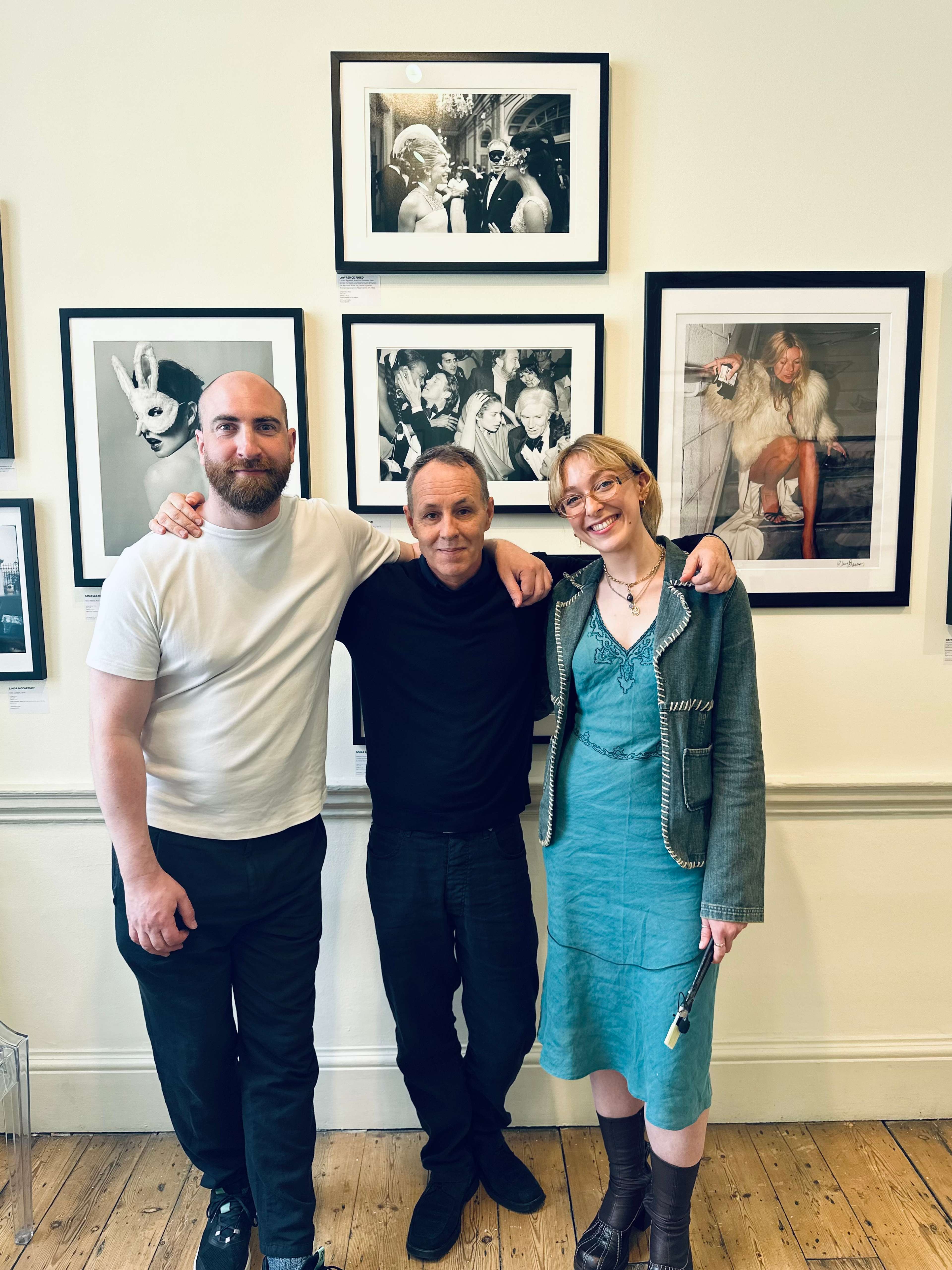 A photograph of Charles Moriarty, Greg Brennan, and Erin-Atlanta Argun at the Iconic Images booth at Photo London, standing in front of photographs hanging on the wall