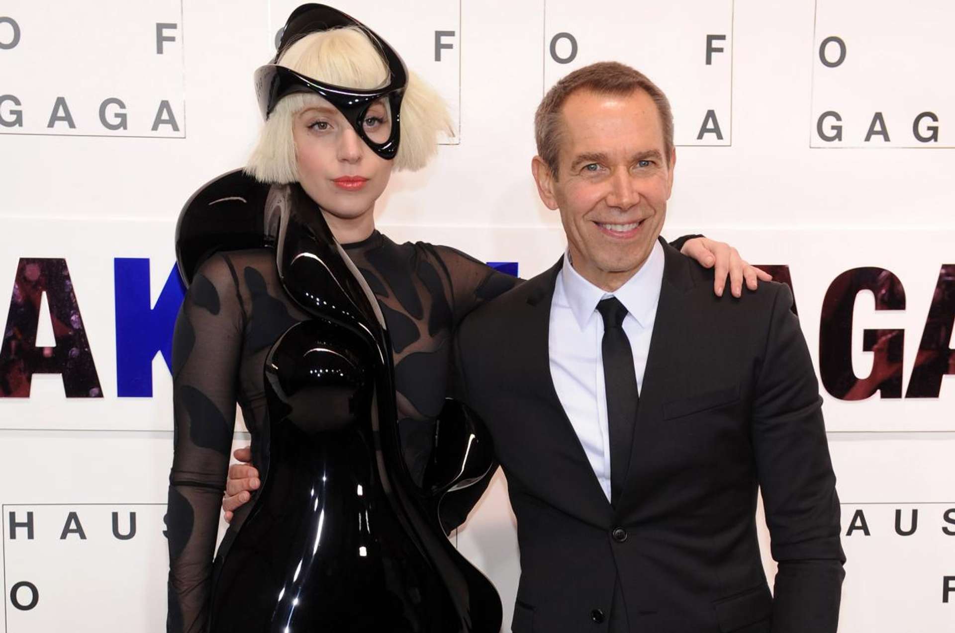 A photograph of artist Jeff Koons alongside musician Lady Gaga. They are embracing against a white background.