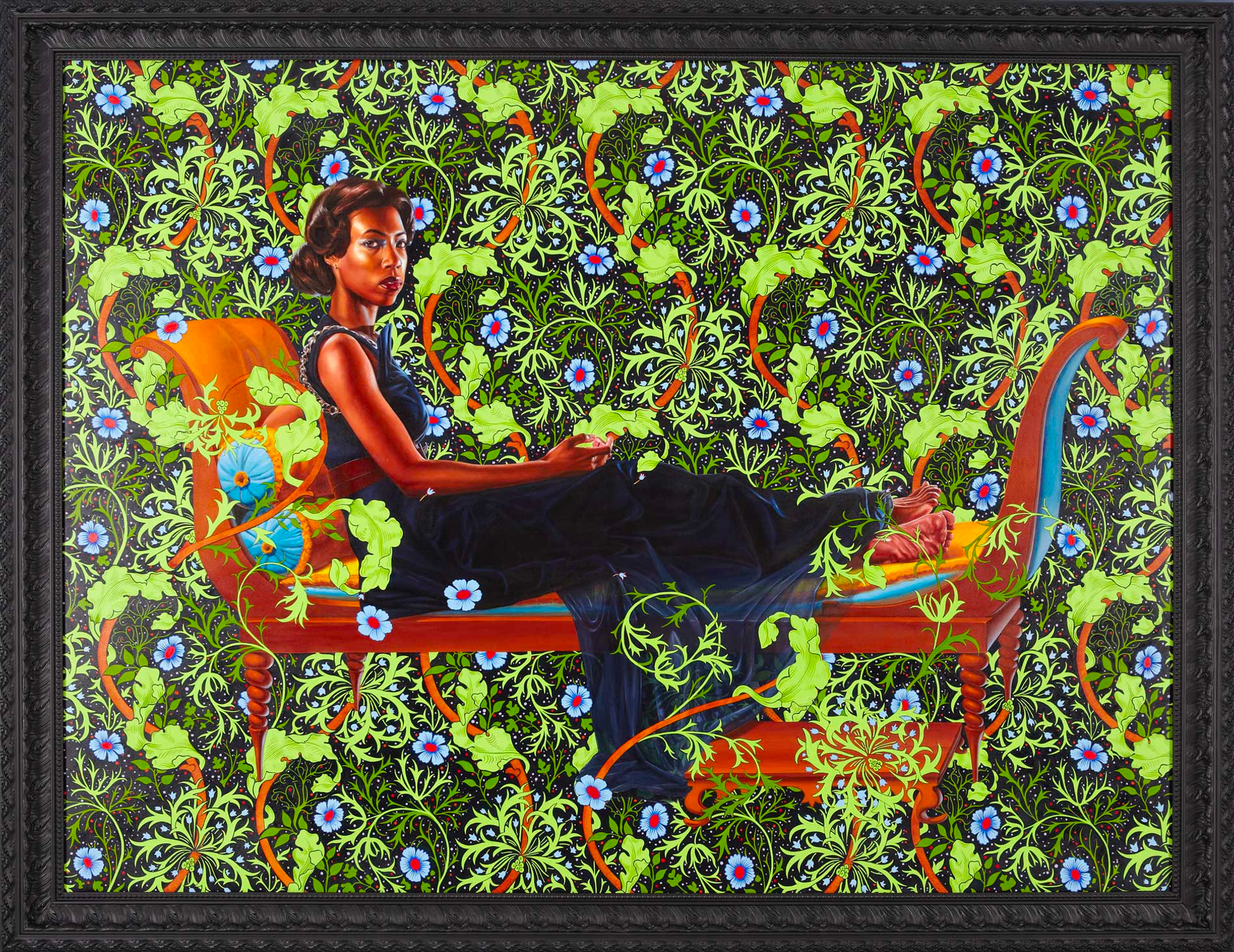 A black woman in a navy blue ball gown sits on a golden brown chaise lounge with turquoise blue cushions. She makes eye contact with the viewer against a vibrant green foliage background adorned with blue flowers.