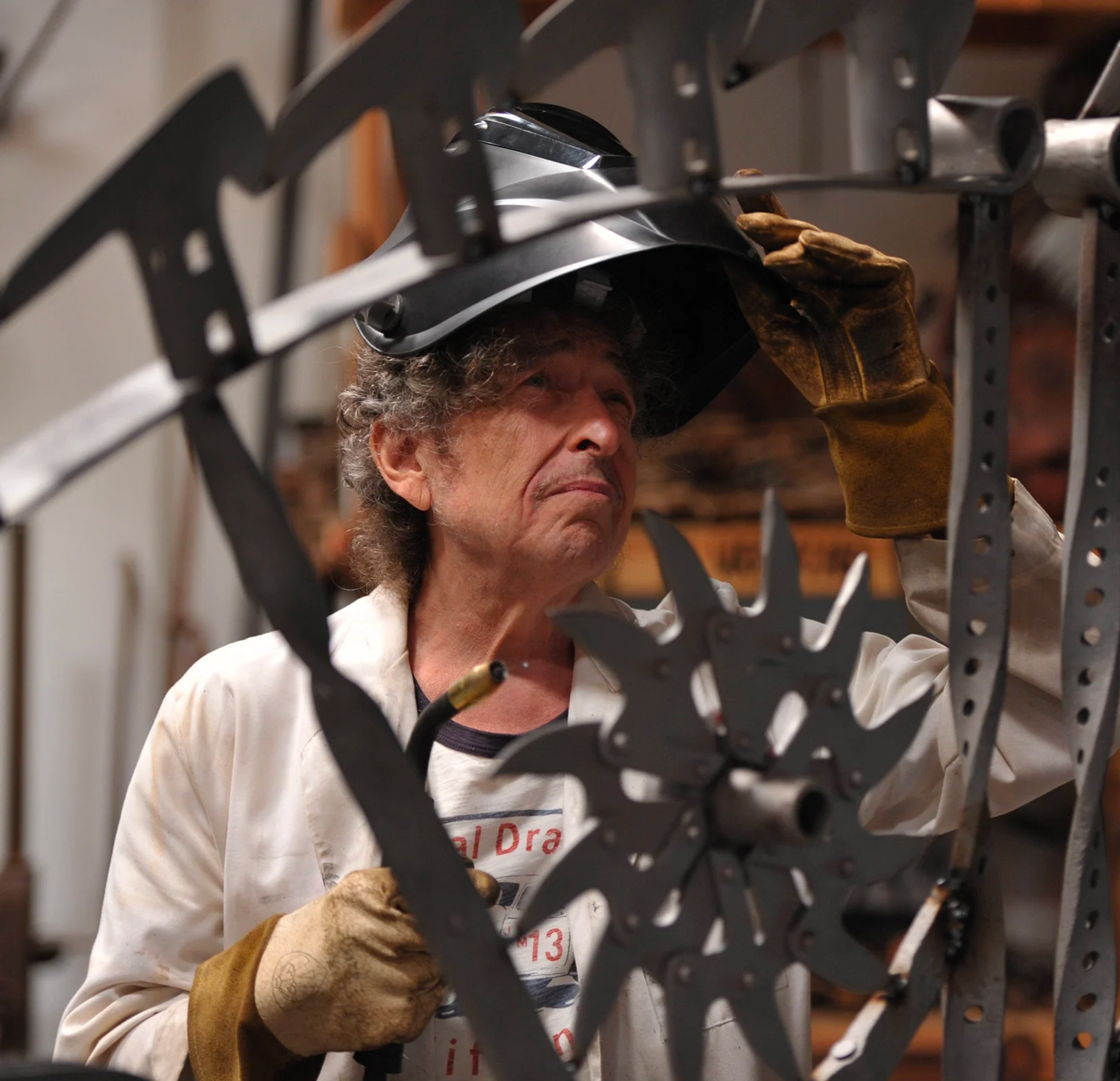 A photographic image of Bob Dyaln wearing yellow gloves and a hardhat working in the studio on a meatl sculpture.