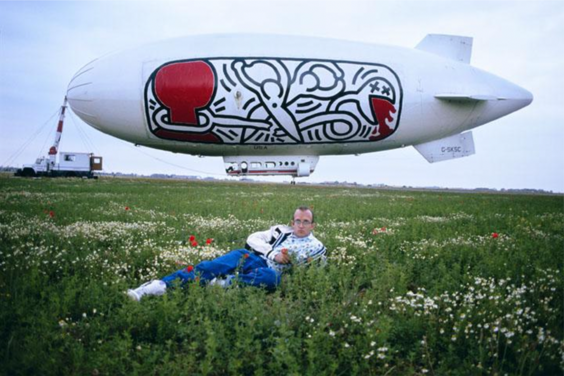 Keith Haring laying on a field of grass with a blimp designed with his artwork behind him in France.