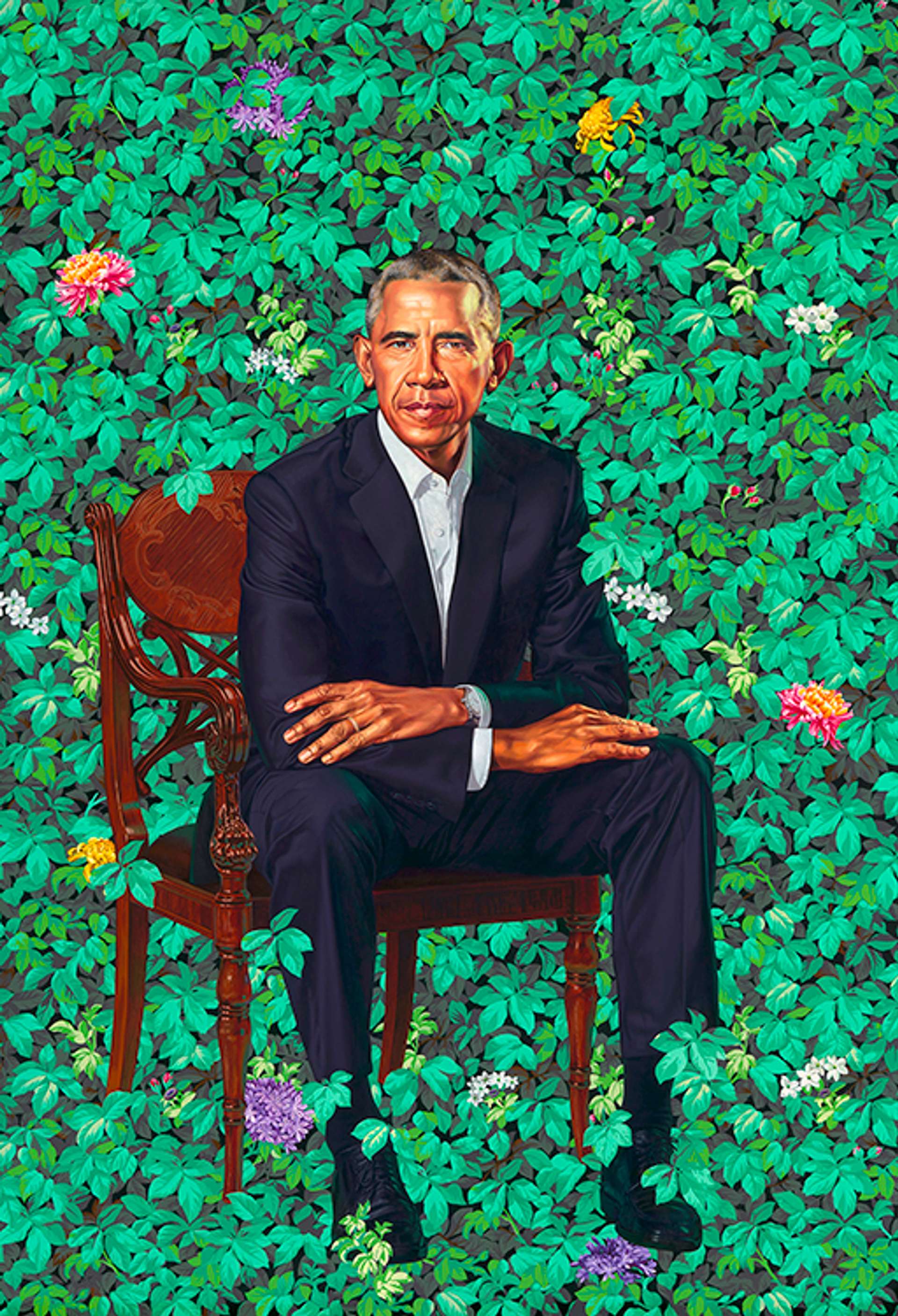 President Barack Obama dressed in a navy blue suit and white shirt, sitting on a wooden chair with his hands in his lap. Lush green leaves and colourful flora populate the background.