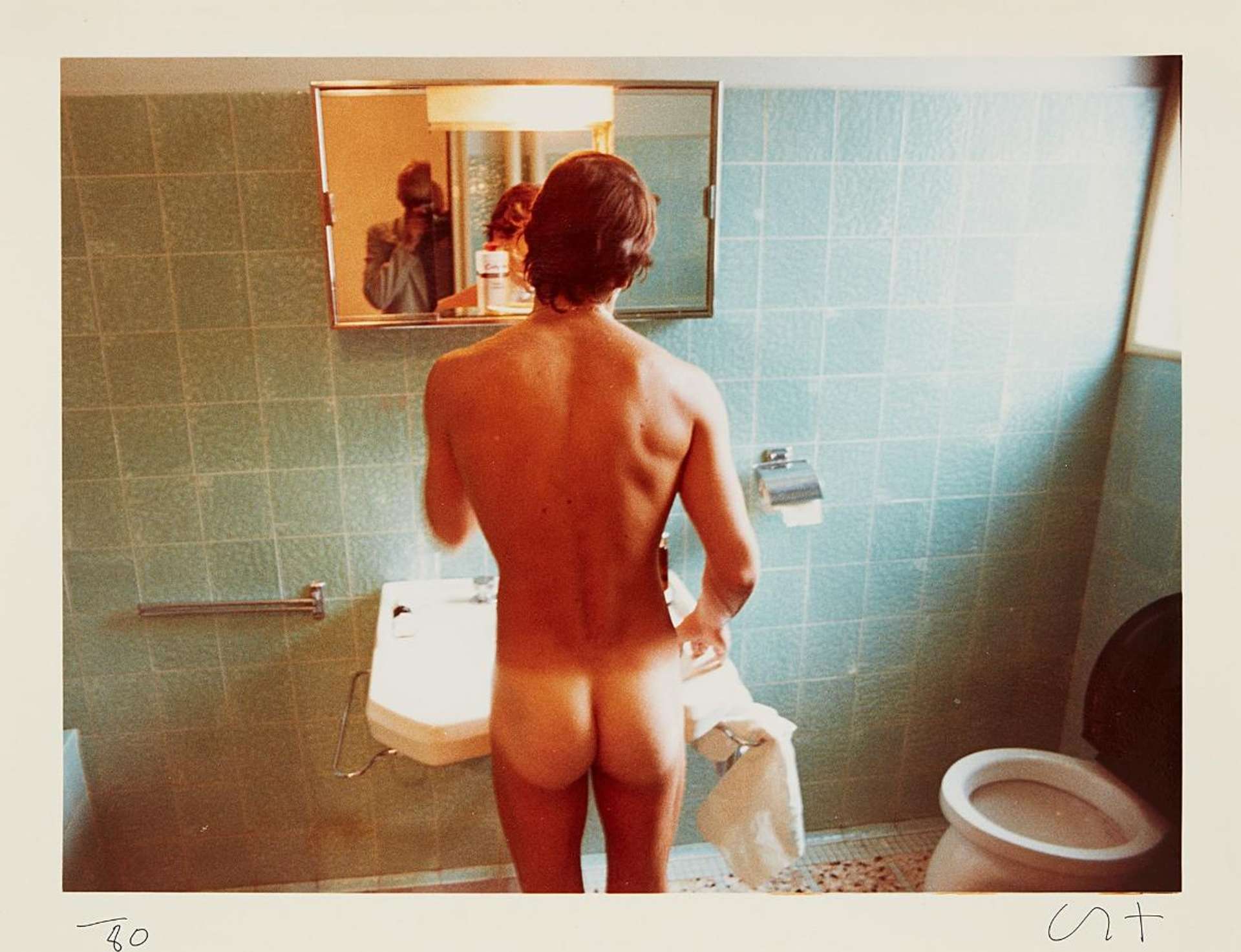 David Hockney’s Peter Washing, Belgrade, September. A digital print of a nude man standing in front of a mirror inside his blue, tiled bathroom. David Hockney is seen in the reflection of the mirror taking the photograph.