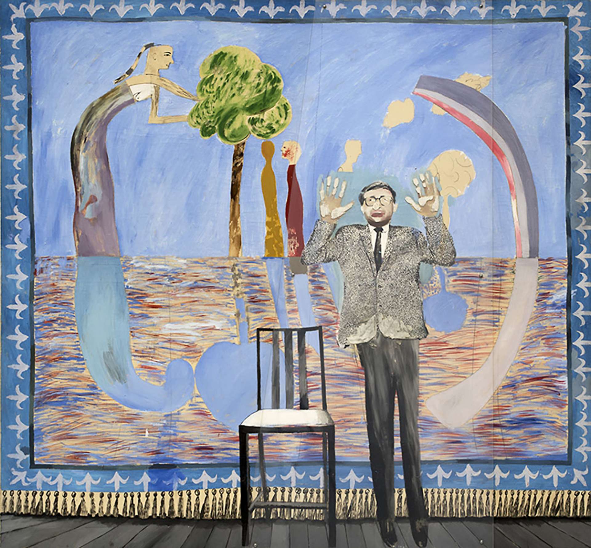 A man stood next to a chair with his hands raised and a decorative tapestry in the background depicting a large figure reaching over a tree