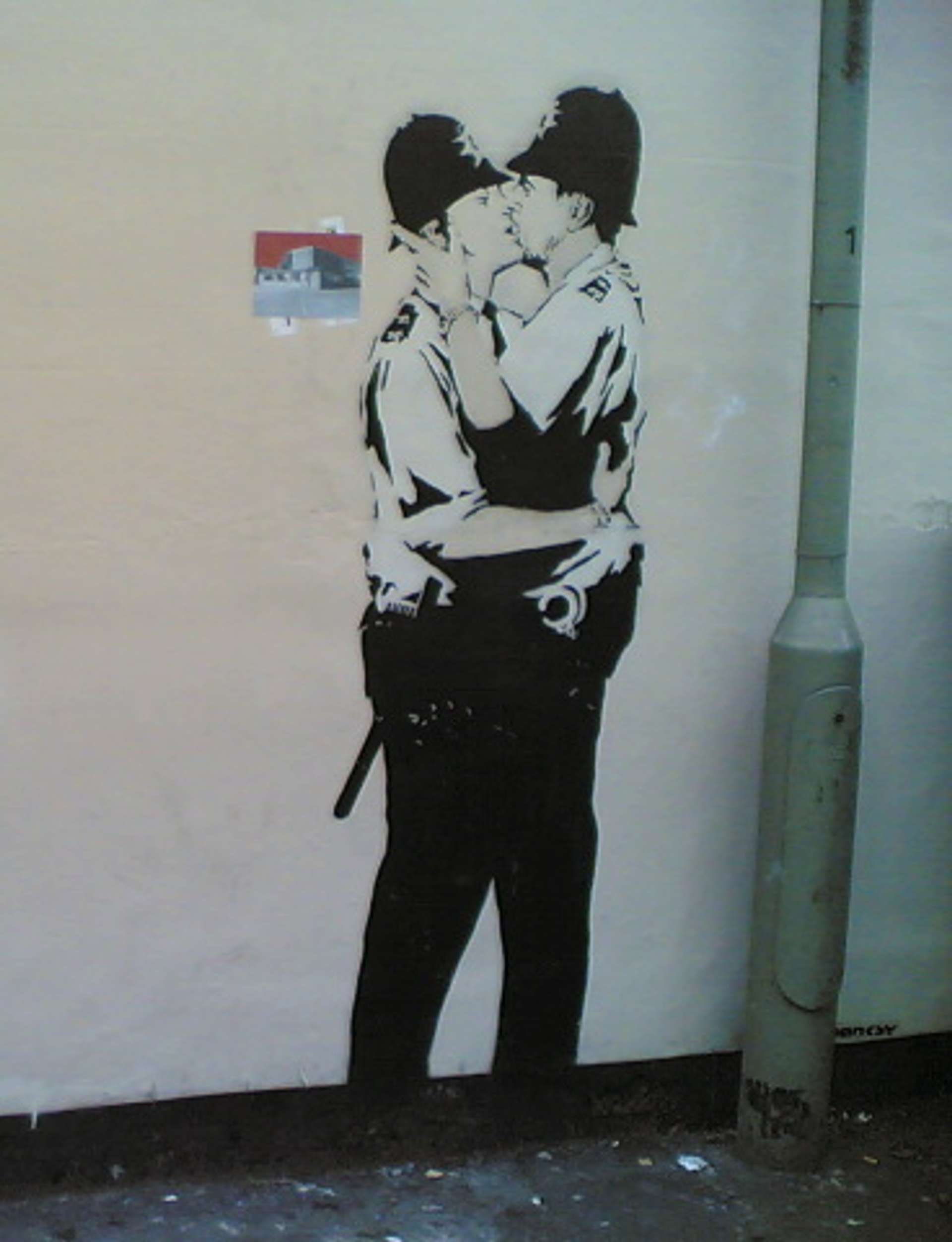 An image of the mural Kissing Coppers by Banksy. It shows two uniformed police officers locked in a passionate kiss.