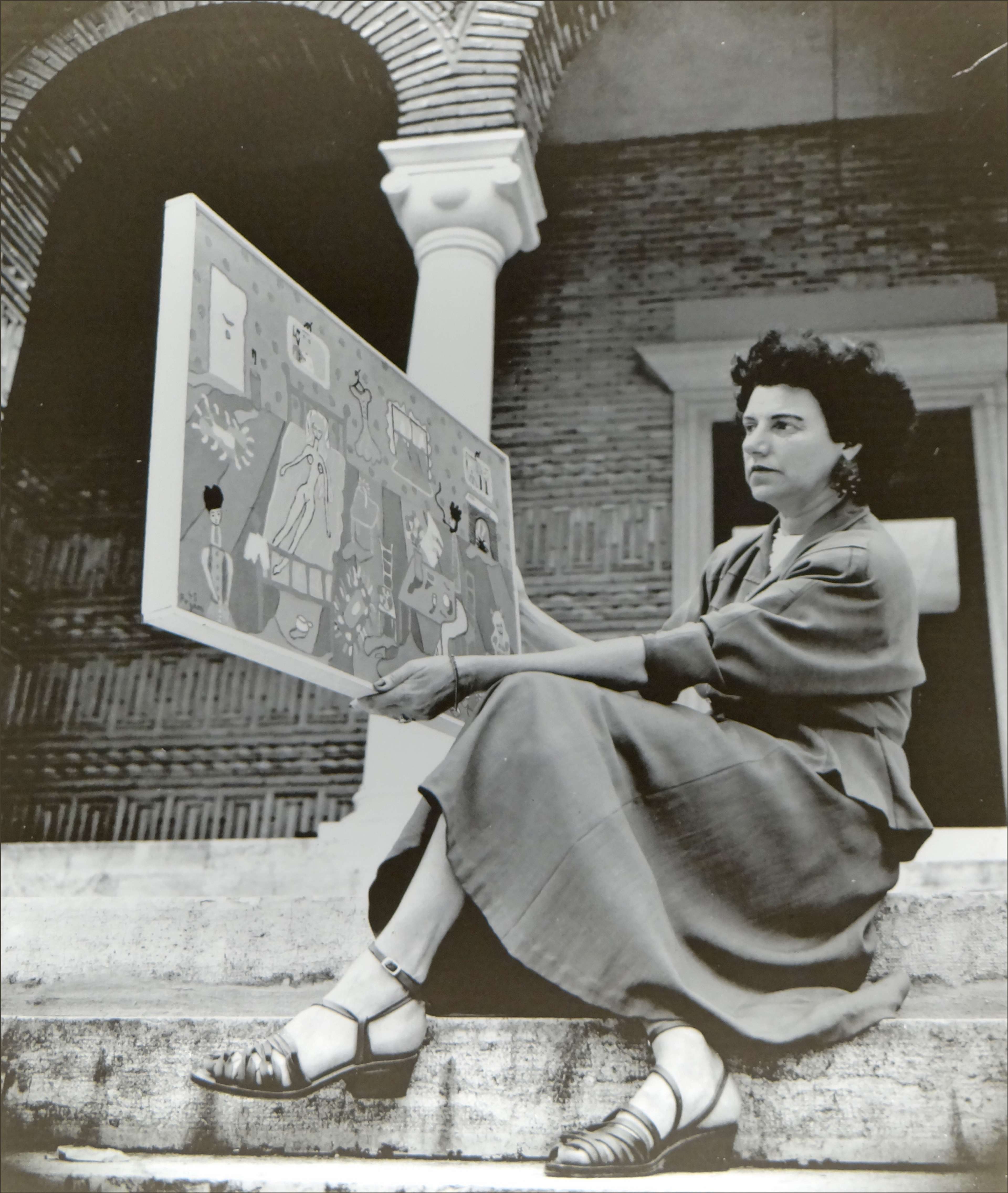 A black and white photograph of a mid-20th century woman sitting and holding up a piece of art