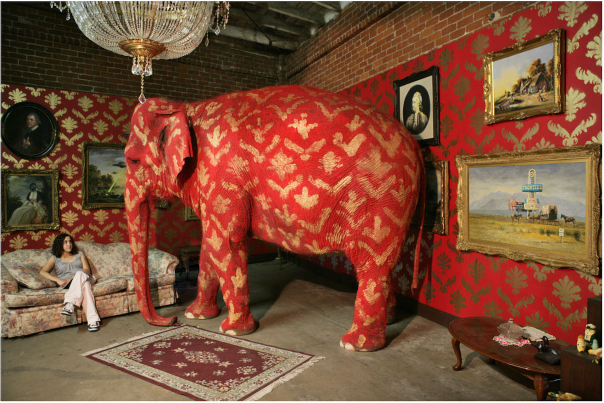 A photo from Banksy's Barely Legal exhibition, capturing a red and gold painted elephant in a room adorned with antique paintings on the walls, an oriental rug, and a couch where a woman is seated, observing the elephant.
