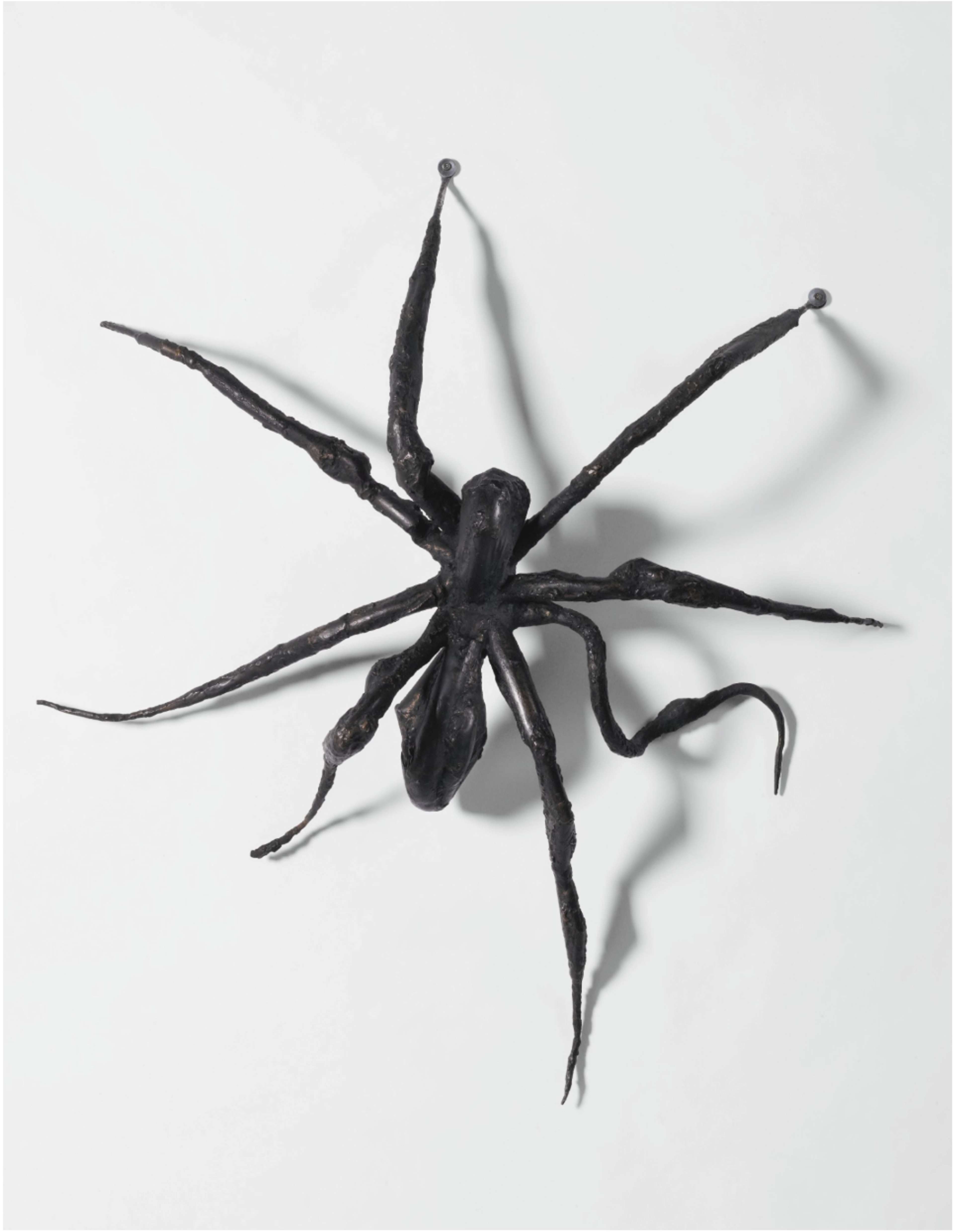 A bronze cast spider sculpture climbing up a white wall, with one leg on the left side slightly bent, creating the illusion of movement.