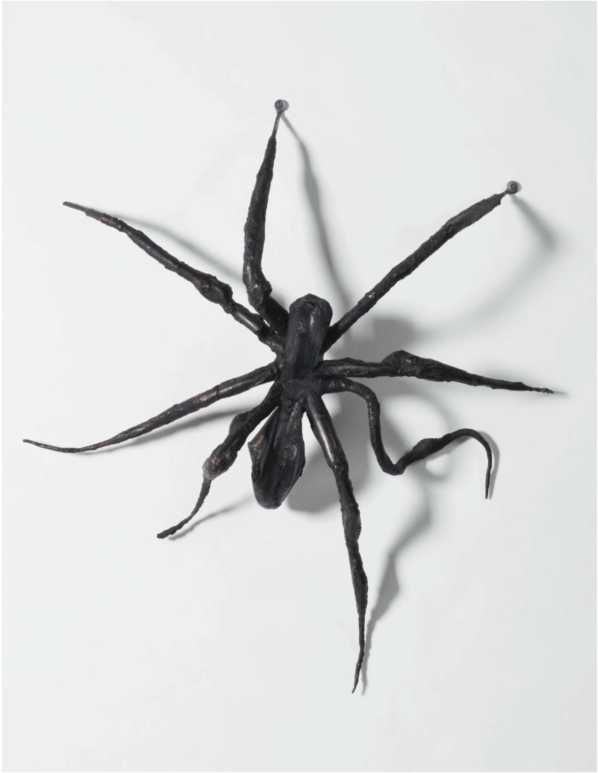 A bronze cast spider sculpture climbing up a white wall, with one leg on the left side slightly bent, creating the illusion of movement.